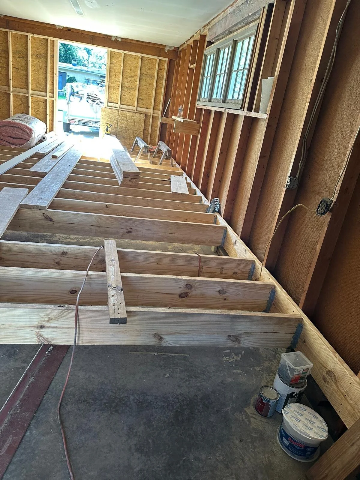 Construction site inside a garage, with wooden framing for a new floor or platform being built, tools, and materials like lumber and paint cans visible.