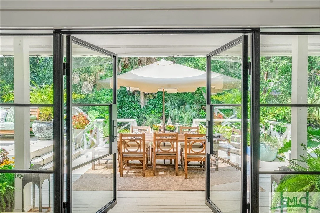 Photo of a screened porch with a dining table and chairs, sun umbrella, surrounded by lush greenery and outdoor furniture.