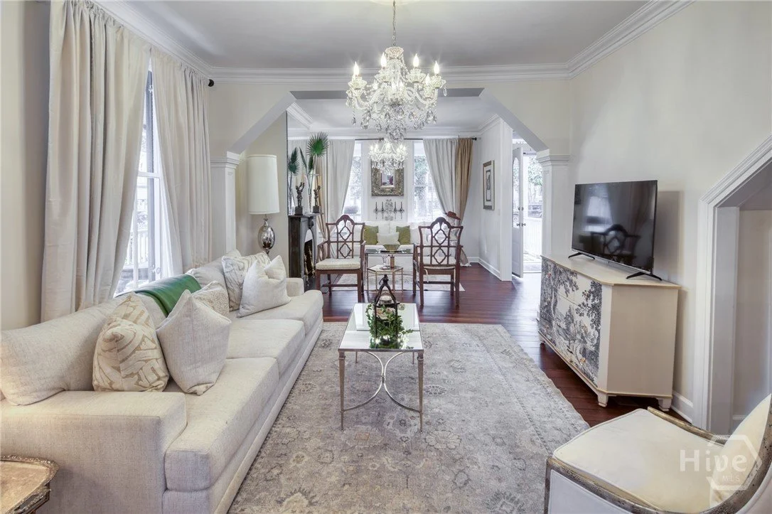 A bright and elegant living room with white walls, curtains, a large chandelier, a beige sofa with pillows, a glass coffee table, a patterned rug, a white TV console, a flat-screen TV, and a dining area with a chandelier, wooden chairs, and a table i