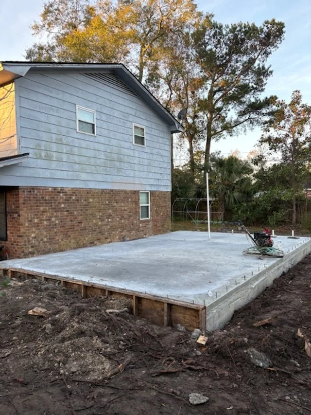 A newly poured concrete patio in the backyard of a house, with construction tools and pipes installed, surrounded by dirt and a wooden form.