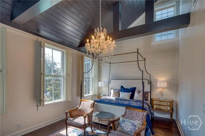 A bedroom with a high, dark vaulted ceiling and a large crystal chandelier. There are two windows with white curtains, a four-poster bed with blue bedding, and two nightstands with lamps. Two upholstered chairs with a small glass-top table are in fro