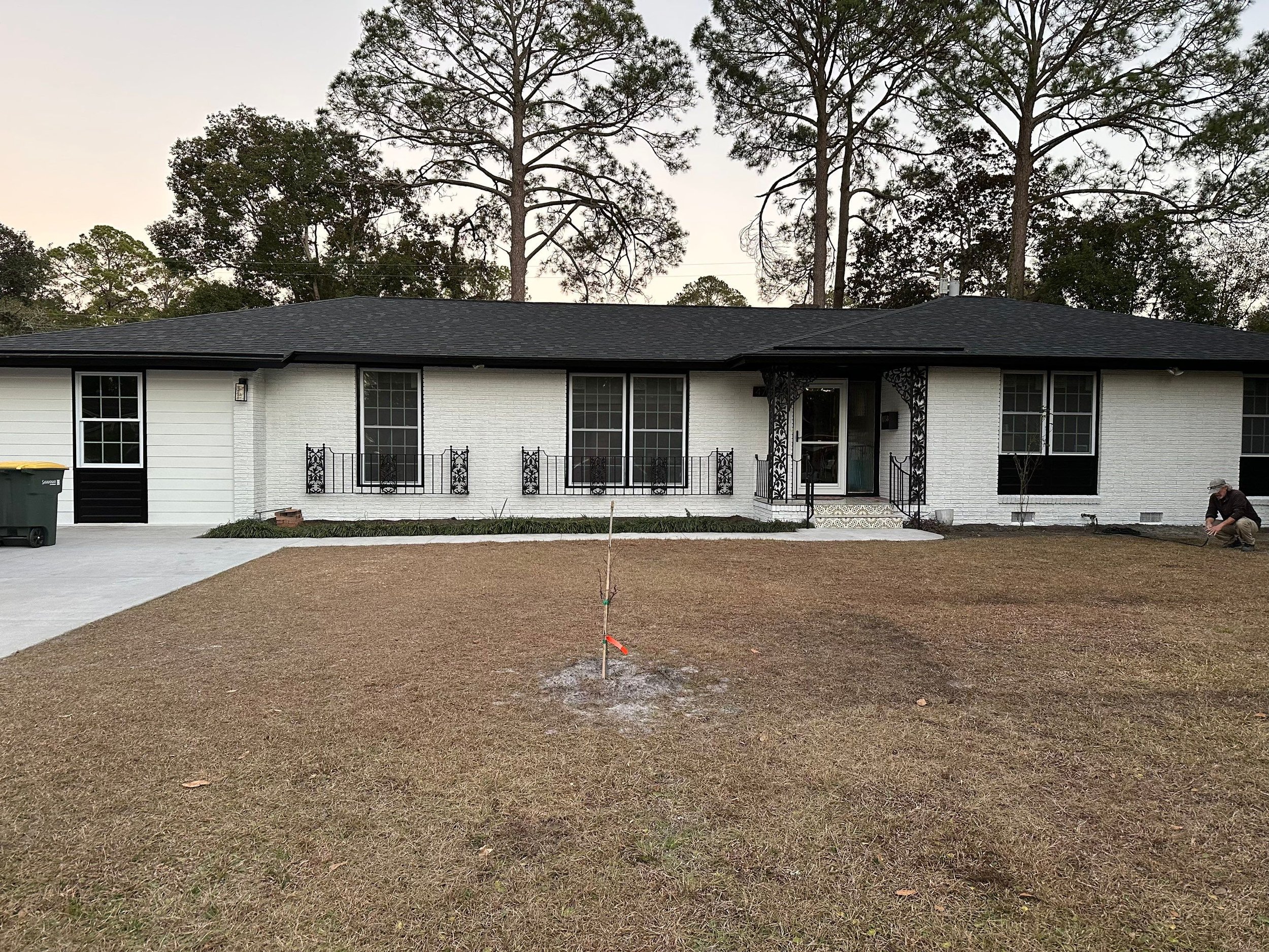 Front view of a newly painted white brick house with black accents, a black roof, and a front lawn with a small tree. A person is seen crouching on the right side planting or inspecting something in the yard. There is a sidewalk leading to the front 