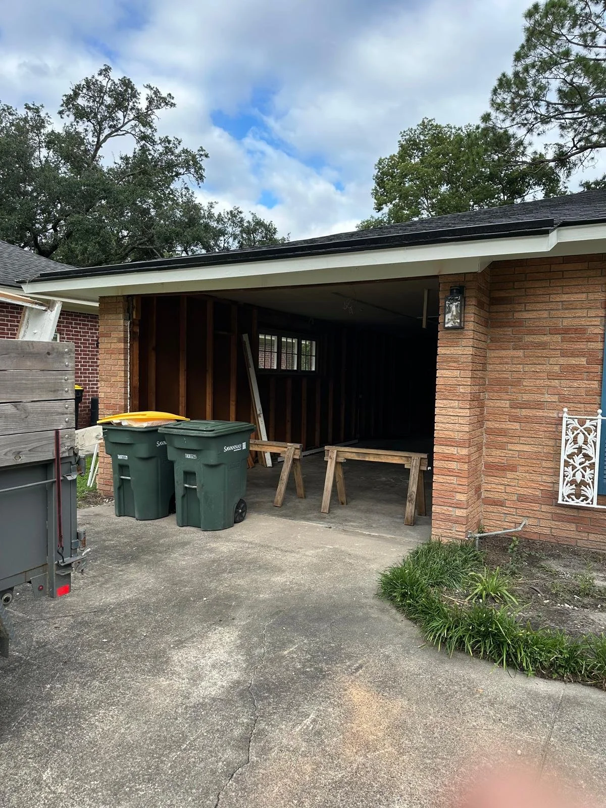 Garage under renovation with wooden framing, five trash bins, a yellow and a white lid, and two wooden sawhorses outside on a concrete driveway, brick house, cloudy sky, and trees in the background.