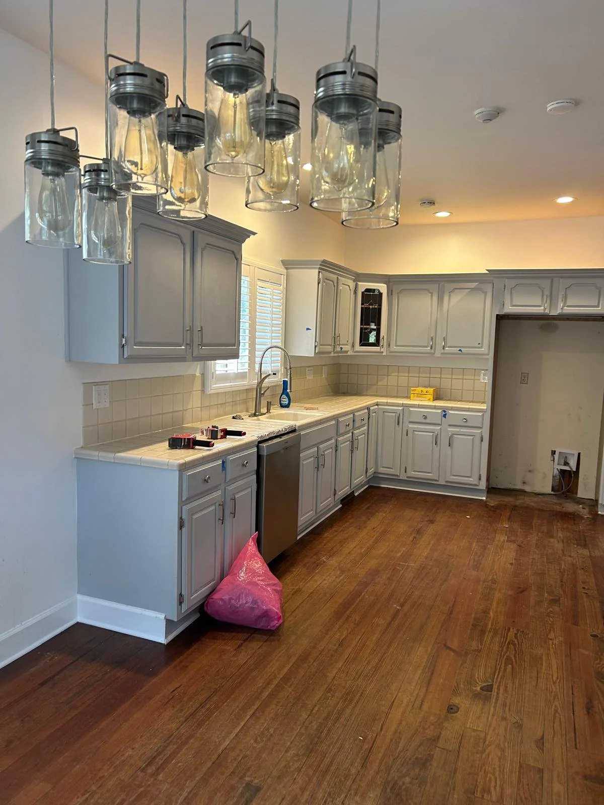 Unfinished kitchen with white cabinets, wooden floors, and a hanging glass light fixture. There are tools on the counter and a pink bag on the floor.