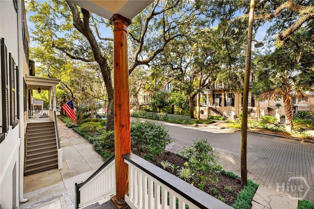 View from a porch showing a tree-lined street with parked cars, greenery, and residential buildings, with an American flag hanging near the steps.