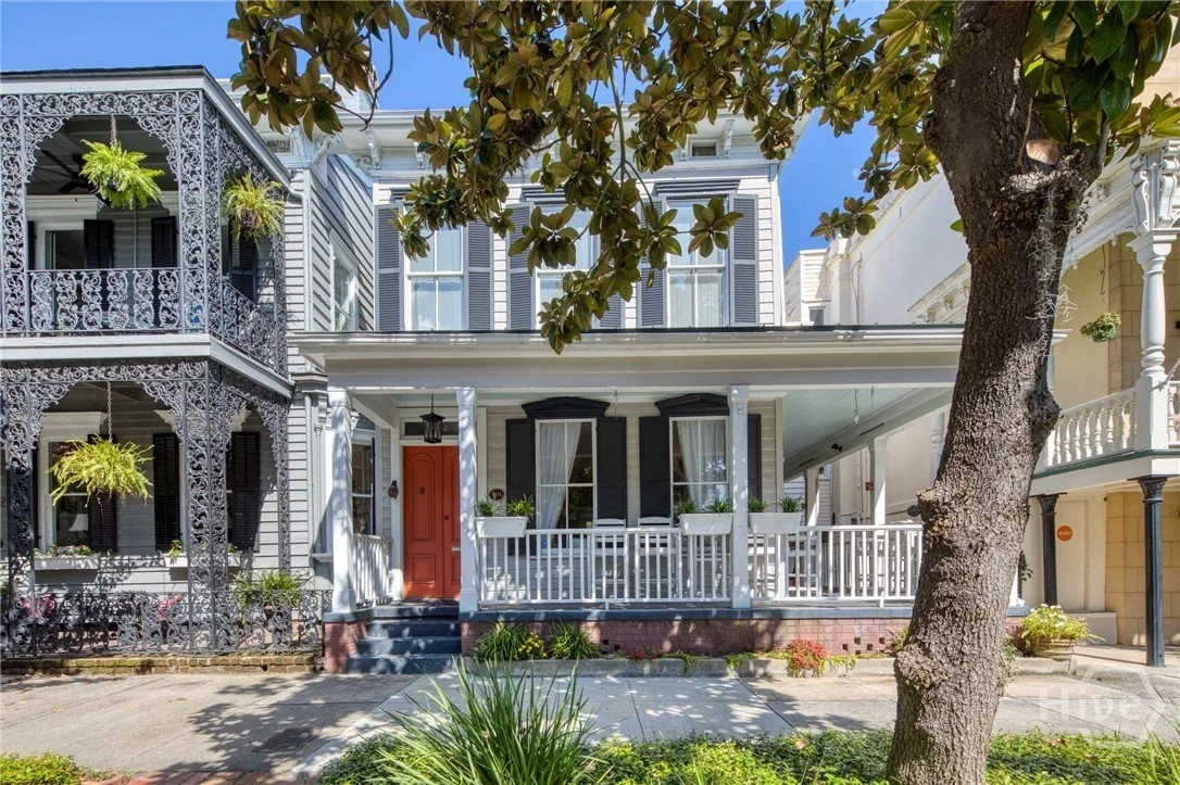 A row of Victorian-style houses with intricate ironwork balconies, a large tree with broad leaves, and a sidewalk in front.