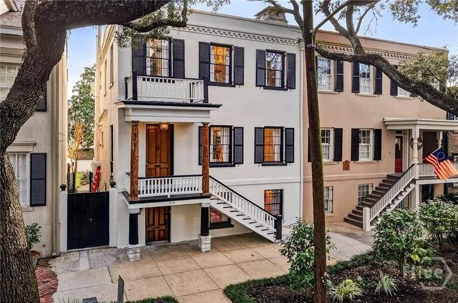 A three-story row house with white exterior, black window shutters, and wooden accents. It has front stairs leading to the second-floor entrance and a small balcony with wrought iron railing. Surrounding buildings and trees are visible.