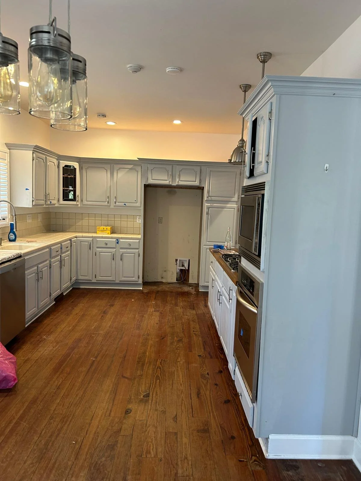 Unfinished kitchen with white cabinets, wooden floor, and appliances, with an empty space in the middle for a stove or refrigerator, and a pink bag in the bottom left corner.