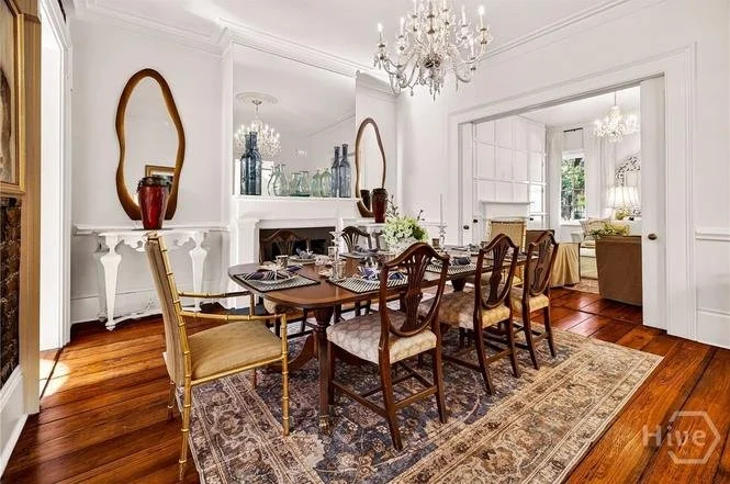 Formal dining room with a wooden table, six upholstered chairs, chandelier lighting, a decorative mirror, a side table with vases, and hardwood floors.