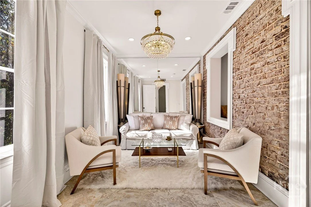 Elegant living room with white sofa and armchairs, brick wall, large windows with white curtains, chandelier, and decorative lighting.