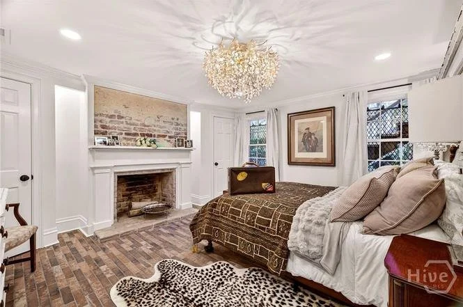 Bedroom with white walls, hardwood floors, and a fireplace with a brick backing. A bed with pillows and a leopard-print rug. A chandelier hanging from the ceiling. Large windows with white curtains and framed artwork on the wall.