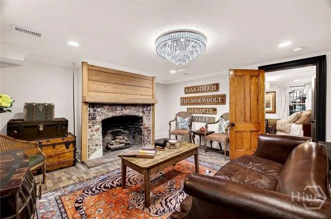 Living room with a brick fireplace, wooden panel above it, chandelier, wooden sign with names, leather chair, vintage trunk, and patterned area rug.