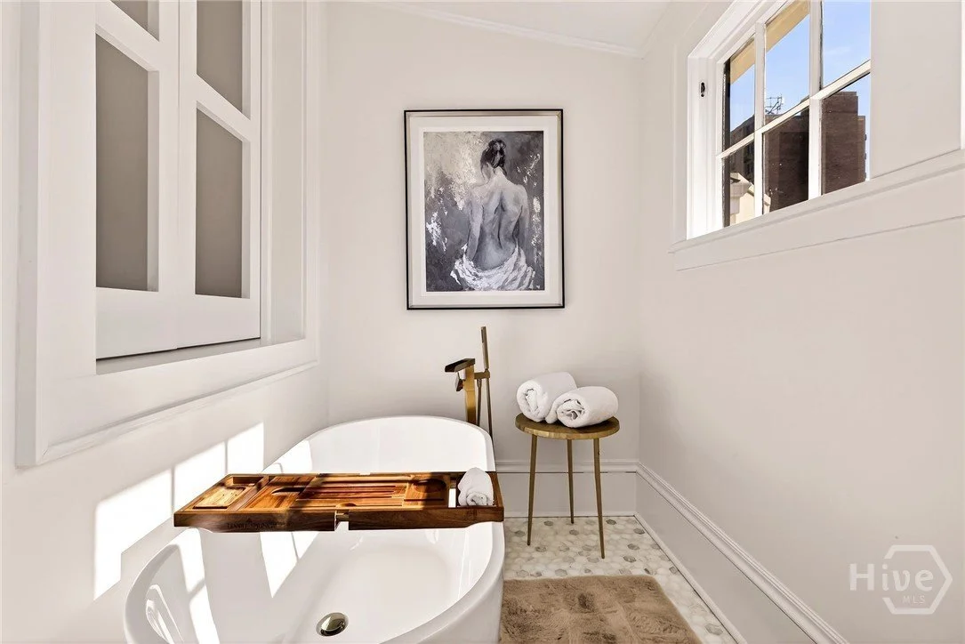 A bathroom with a white bathtub, a wooden bathtub tray, and rolled white towels on a brass side table. There is a black and white artwork of a woman on the wall and a window with sunlight streaming in.