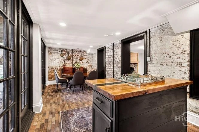 Open-concept dining and kitchen area with exposed brick walls, hardwood flooring, black window frames, a wooden dining table, upholstered chairs, and a kitchen island with a wooden countertop.