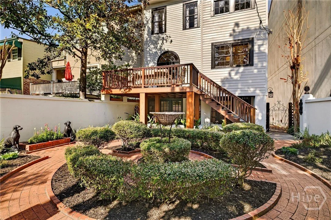 Backyard garden with trimmed bushes, brick pathway, decorative dog statues, a wooden deck with stairs, and a white house with black-framed windows.