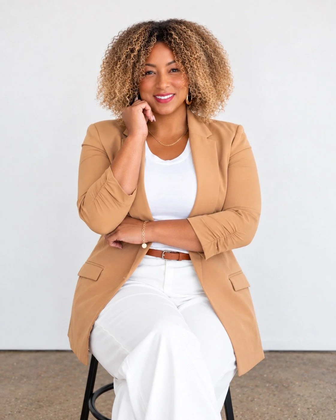 A woman with curly blonde hair wearing a tan blazer, white shirt, and white pants sitting on a black chair against a plain white background.