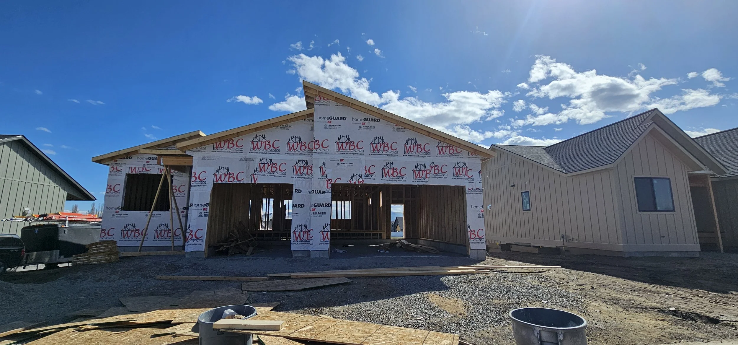 House under construction with wooden framing and exterior wall covering, set against blue sky with scattered clouds.