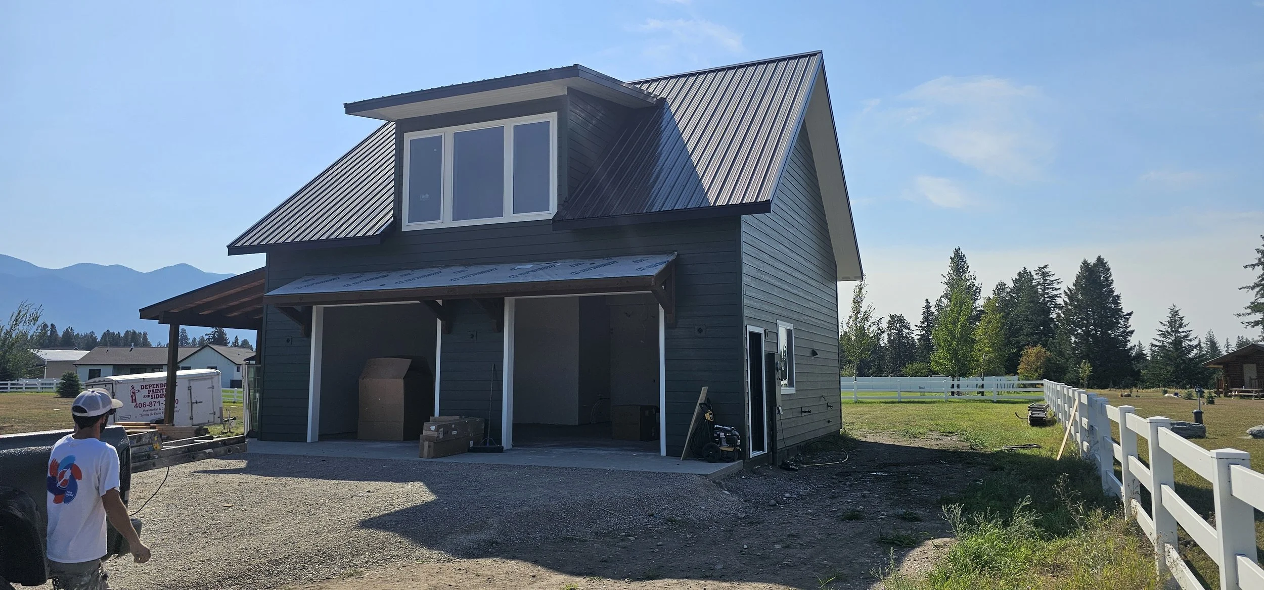 A two-story house under construction with gray siding and a metal roof, surrounded by a gravel driveway and a white fence, with mountains and trees in the background on a sunny day.