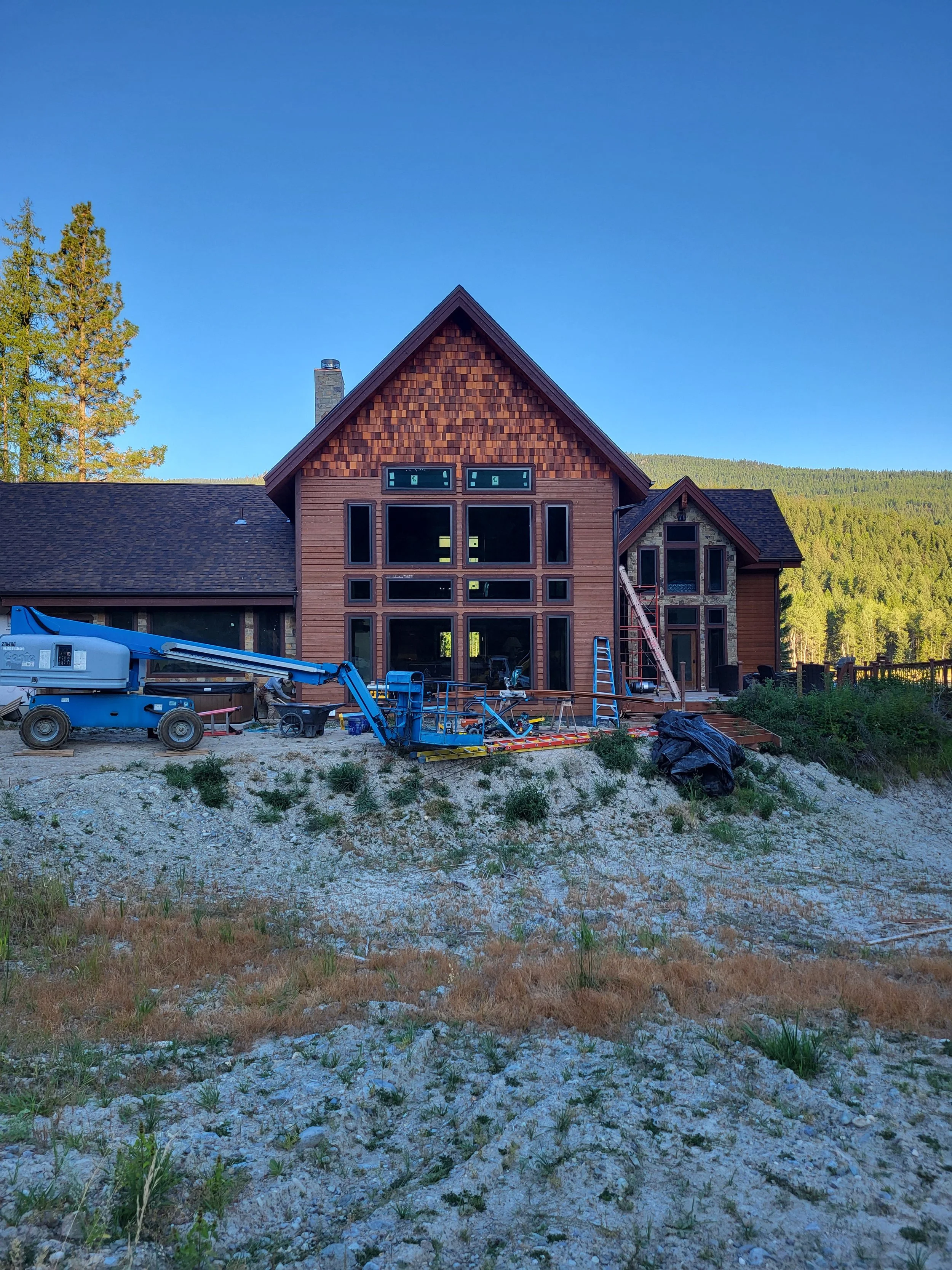 A house under construction with a wooden exterior, large windows, and construction equipment in the front yard on a dirt and gravel surface, surrounded by trees and mountains in the background.