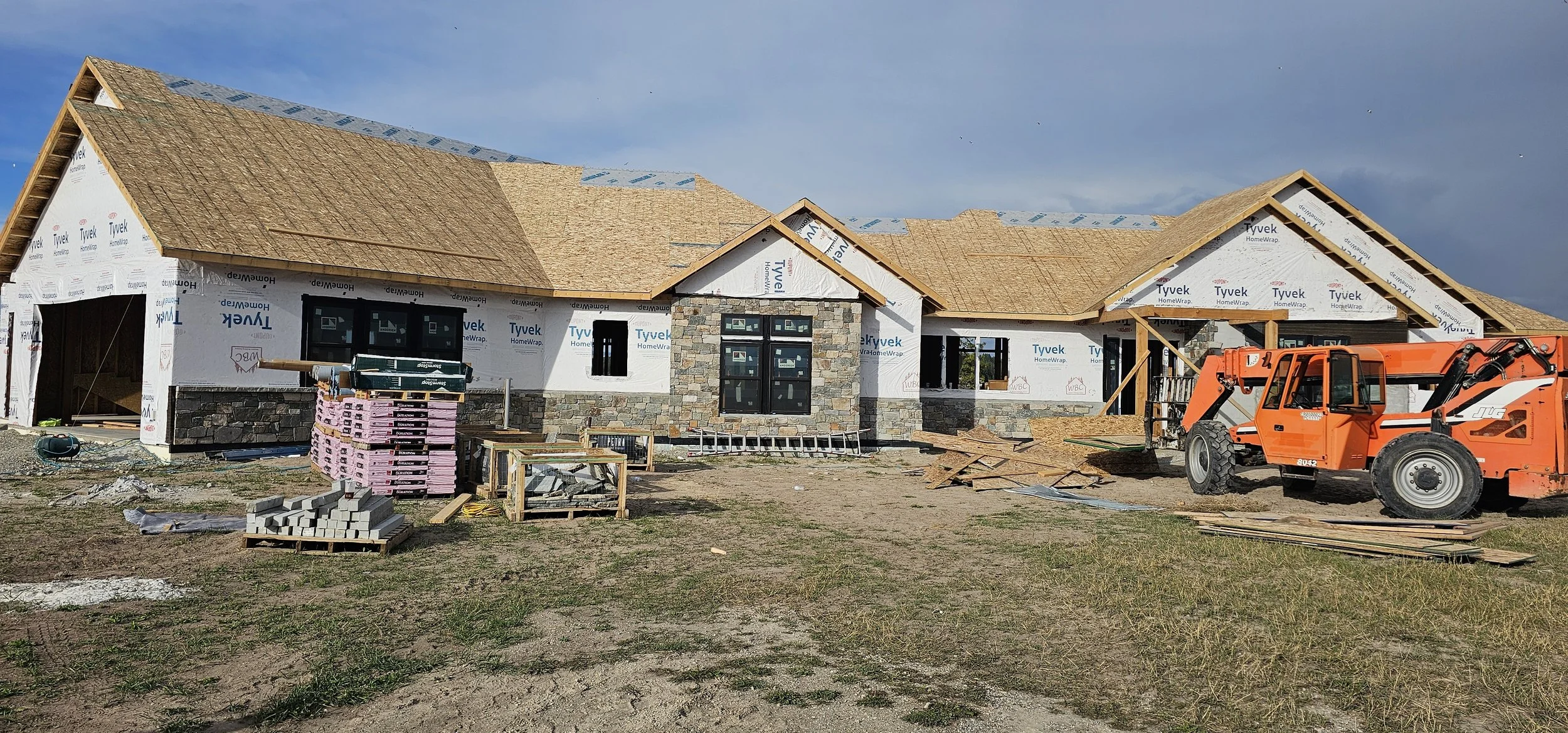 A house under construction with a partially built wooden roof, stone accents on the front, and construction materials and machinery around the site.