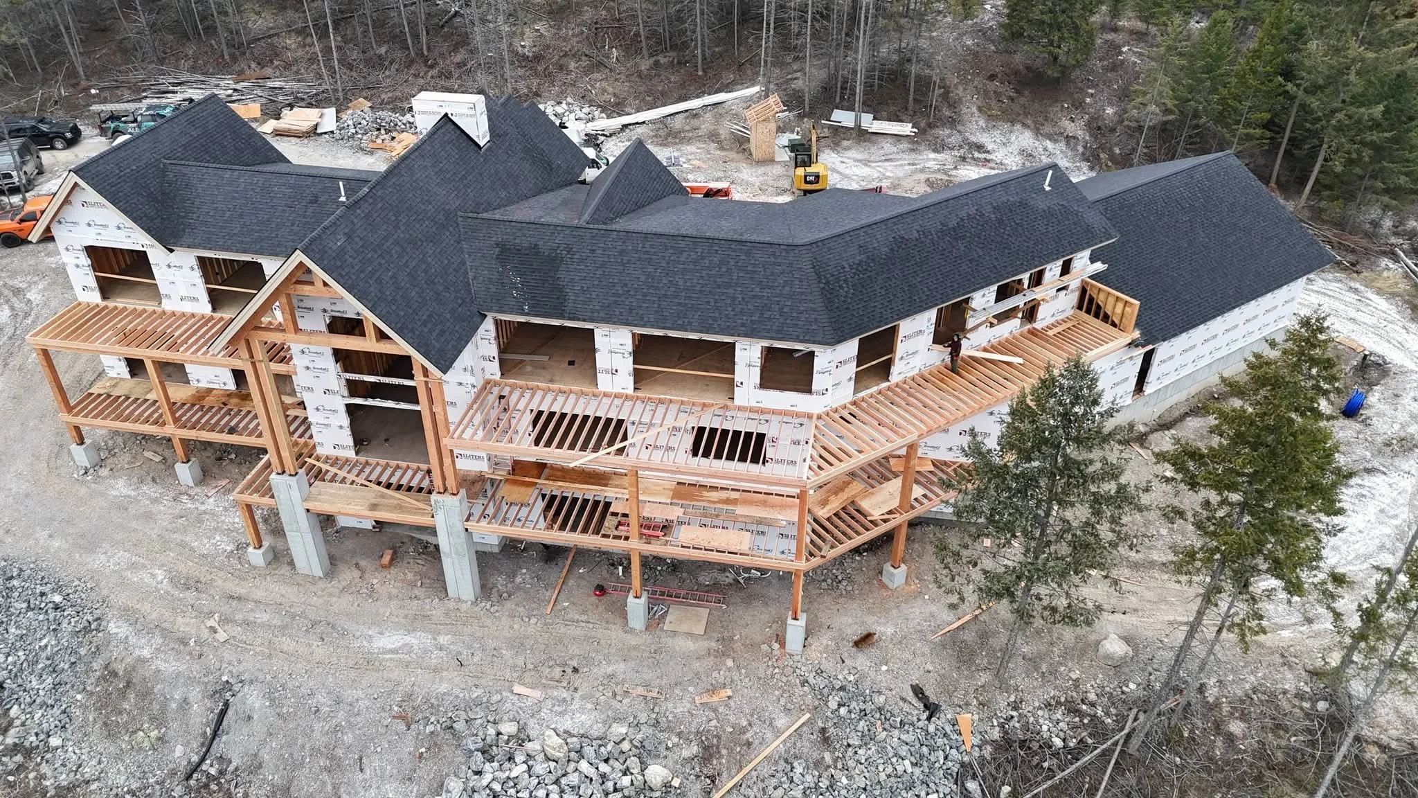 A large house under construction with wooden framework and a dark shingled roof, situated on a rocky and wooded lot.