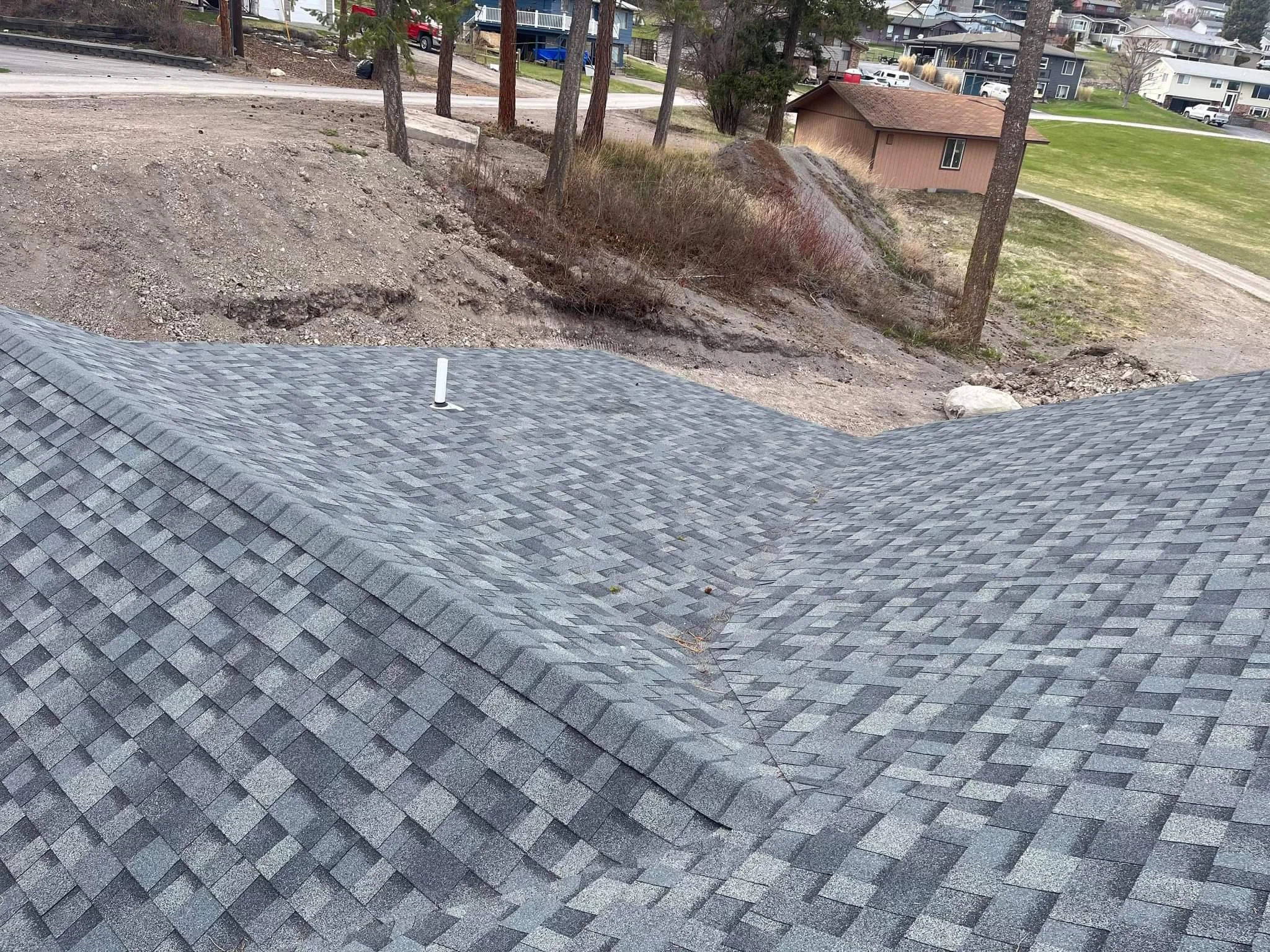 Photo shows a rooftop with gray asphalt shingles, a white pipe vent, and a view of a yard with dirt, trees, and a small brown building in the background.
