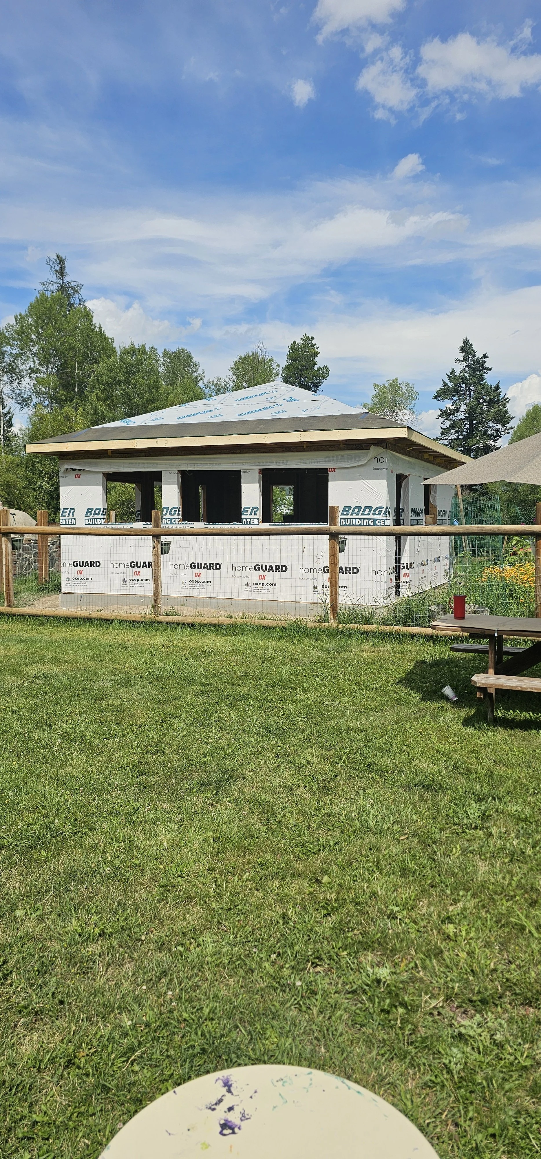 A partially constructed building with a wooden frame and white insulation wrapping, surrounded by a grassy yard with trees in the background and a blue sky with some clouds.