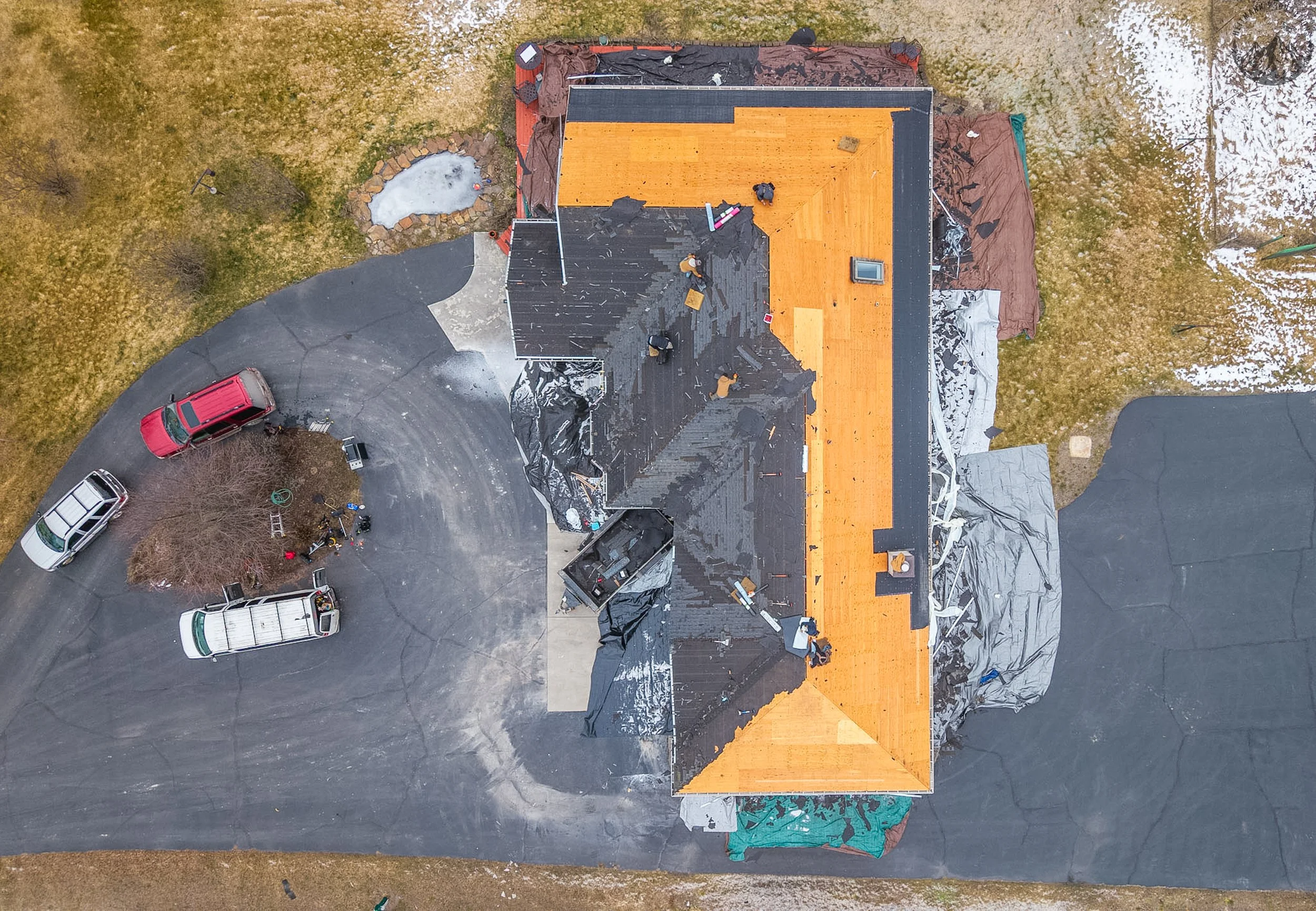An aerial view of a house under construction with roofing work ongoing, surrounded by a driveway and lawn with some snow patches.