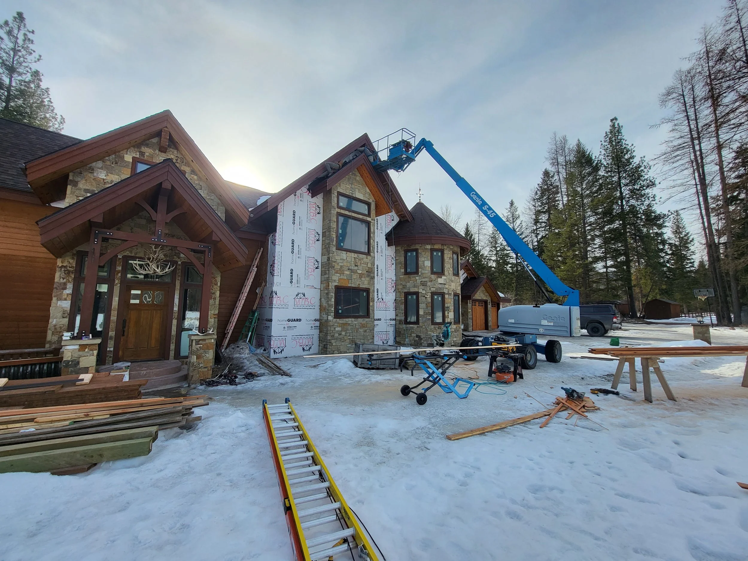 Construction workers use a crane to work on the roof of a large house under construction in a snowy yard surrounded by trees.
