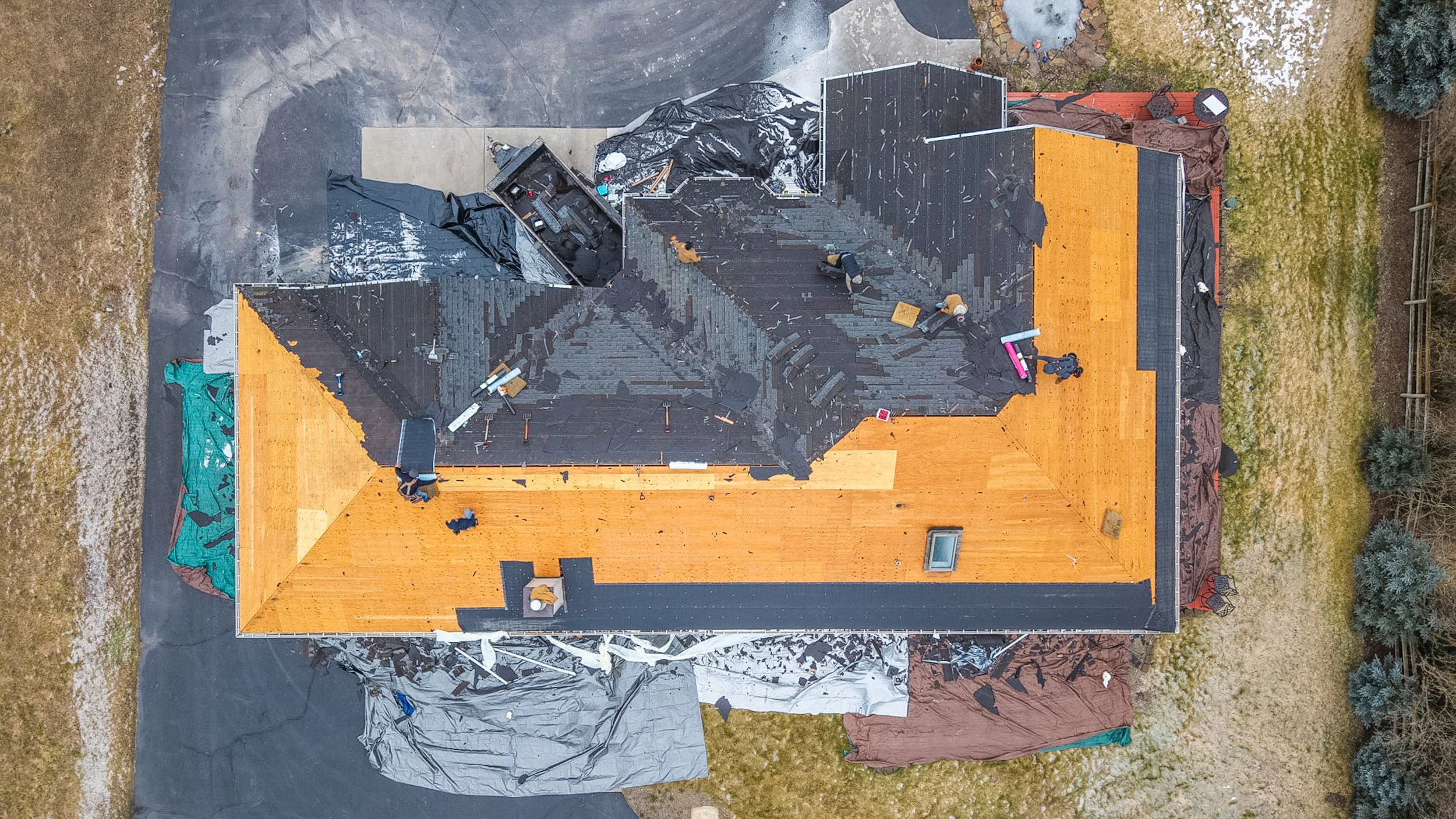 Bird's eye view of a house under renovation, with part of the roof stripped and workers present, surrounded by a yard with grass, trees, and construction materials covered with tarps.
