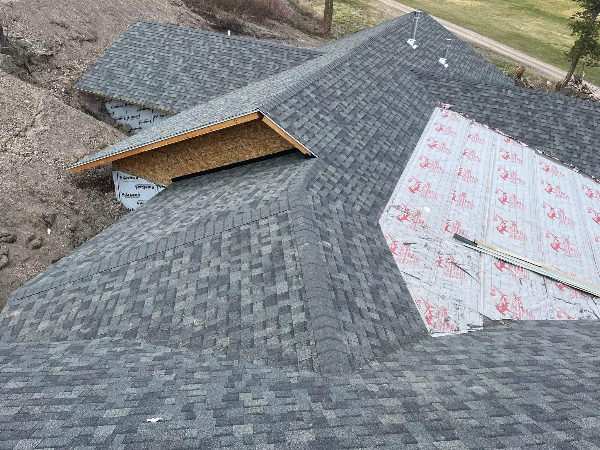 View of a house roof under construction, showing asphalt shingles partly installed, with some sections covered by underlayment and open sheathing, surrounded by dirt and greenery.