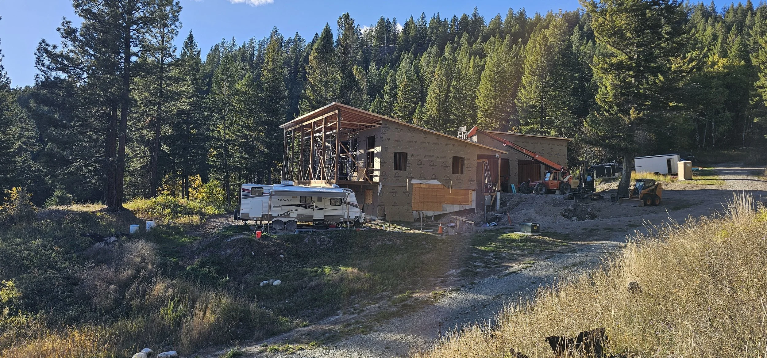 Under construction house in a forested area with construction equipment and a camper trailer.