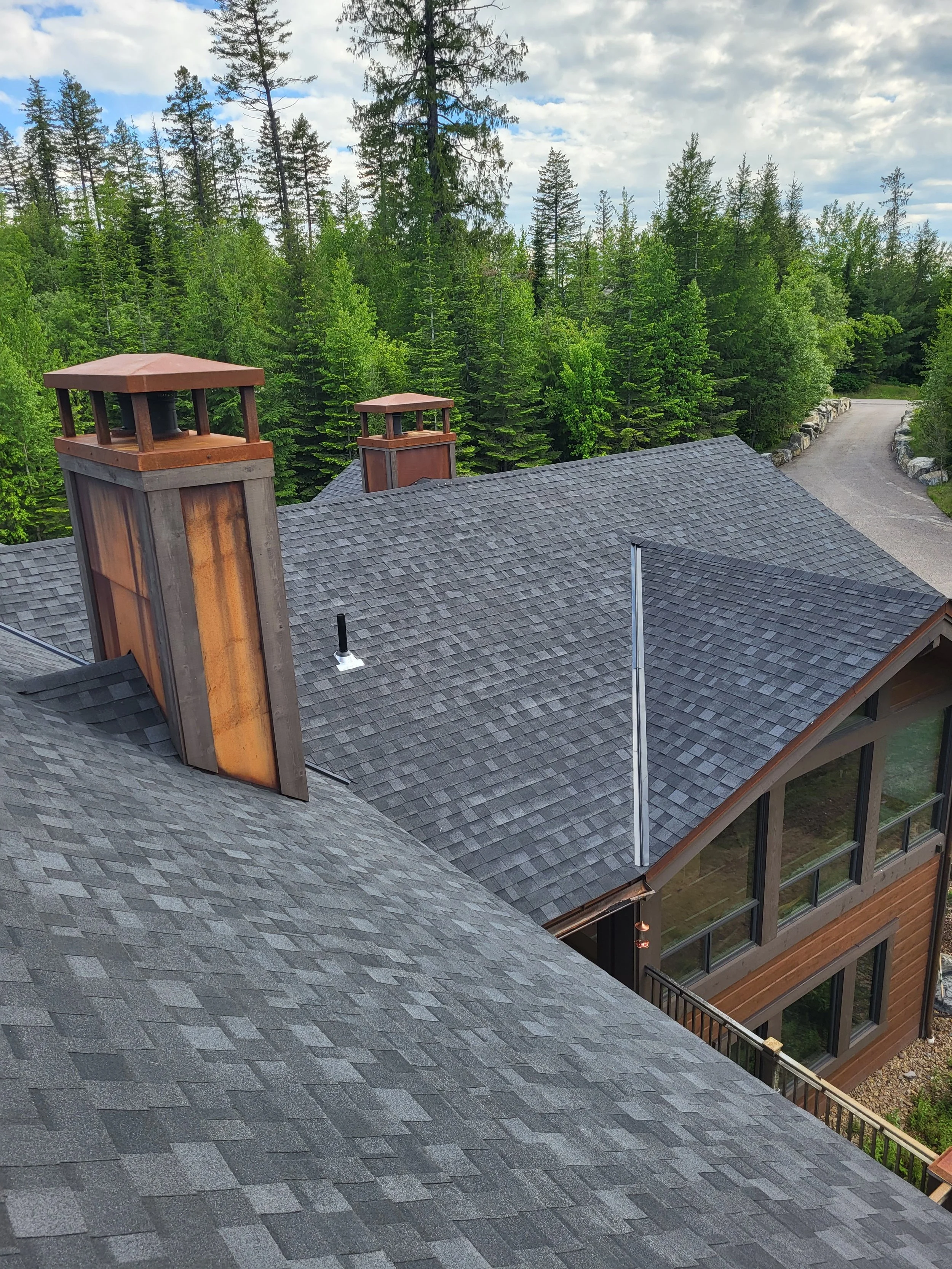 View of a house's gray shingled roof with three brick chimneys, surrounded by a lush green forest and a winding driveway.
