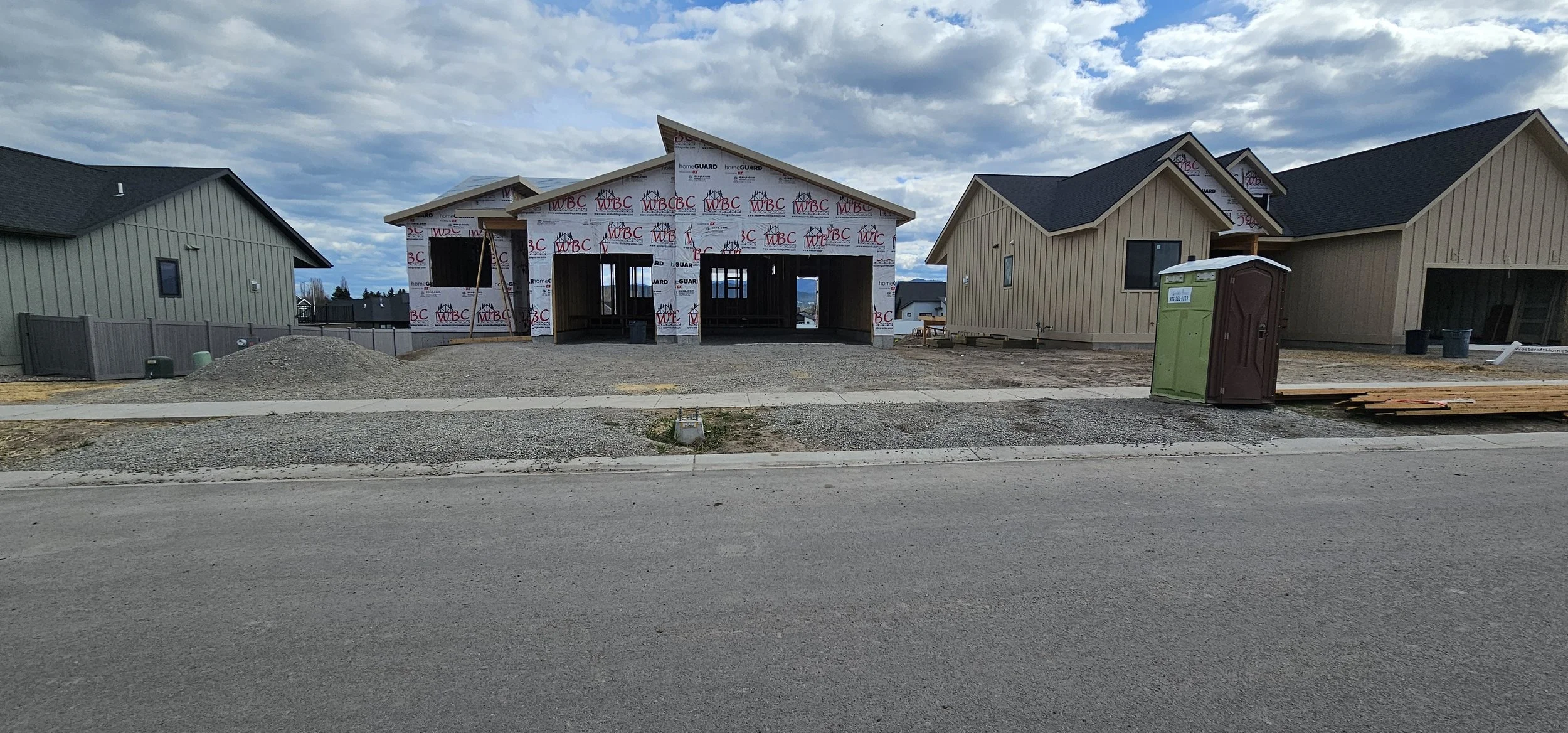 Under construction house in a neighborhood with completed houses on either side, construction materials, a portable toilet, and a partly cloudy sky.