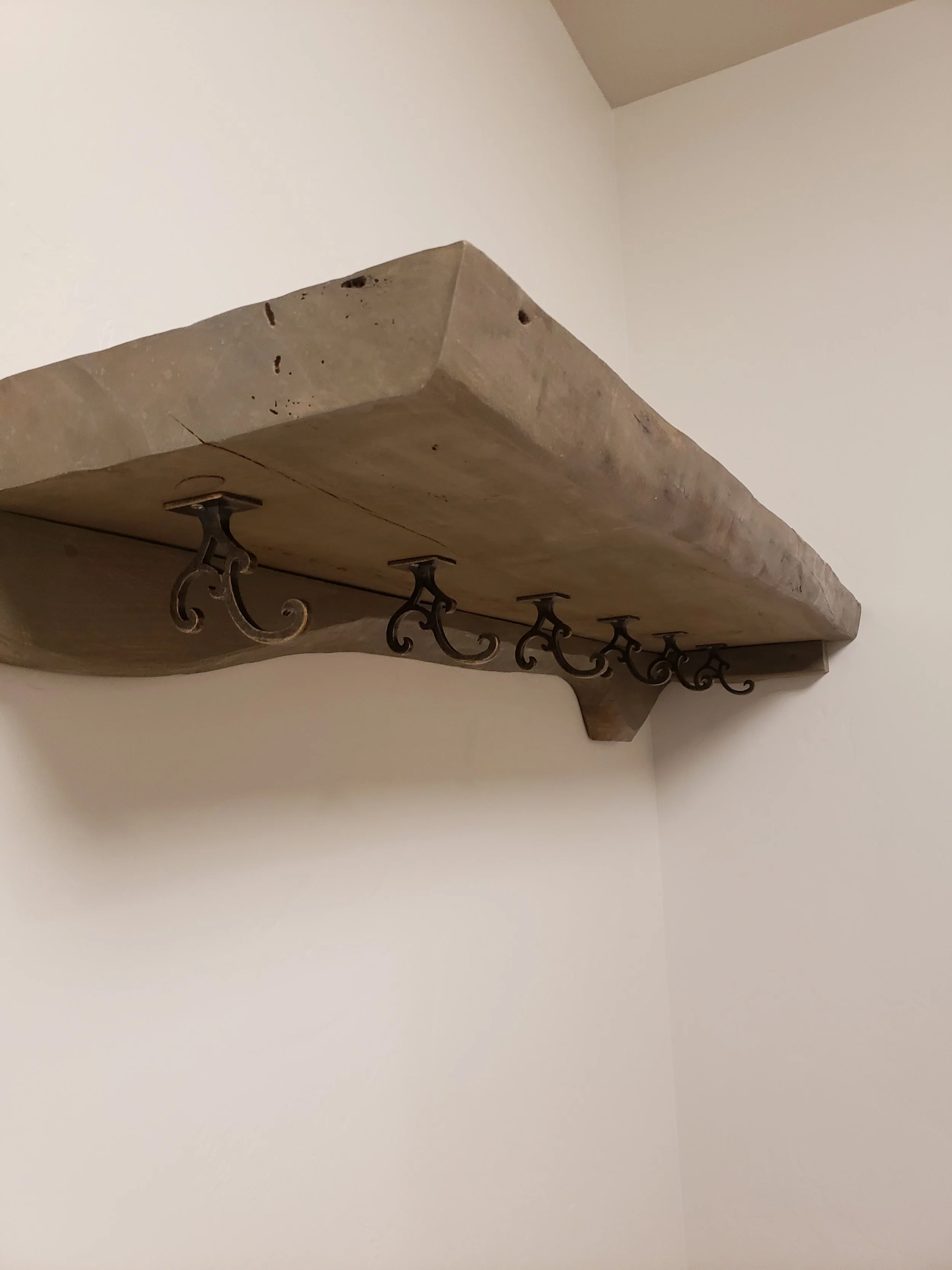 A wooden shelf with decorative iron hooks mounted on a white wall in a corner.