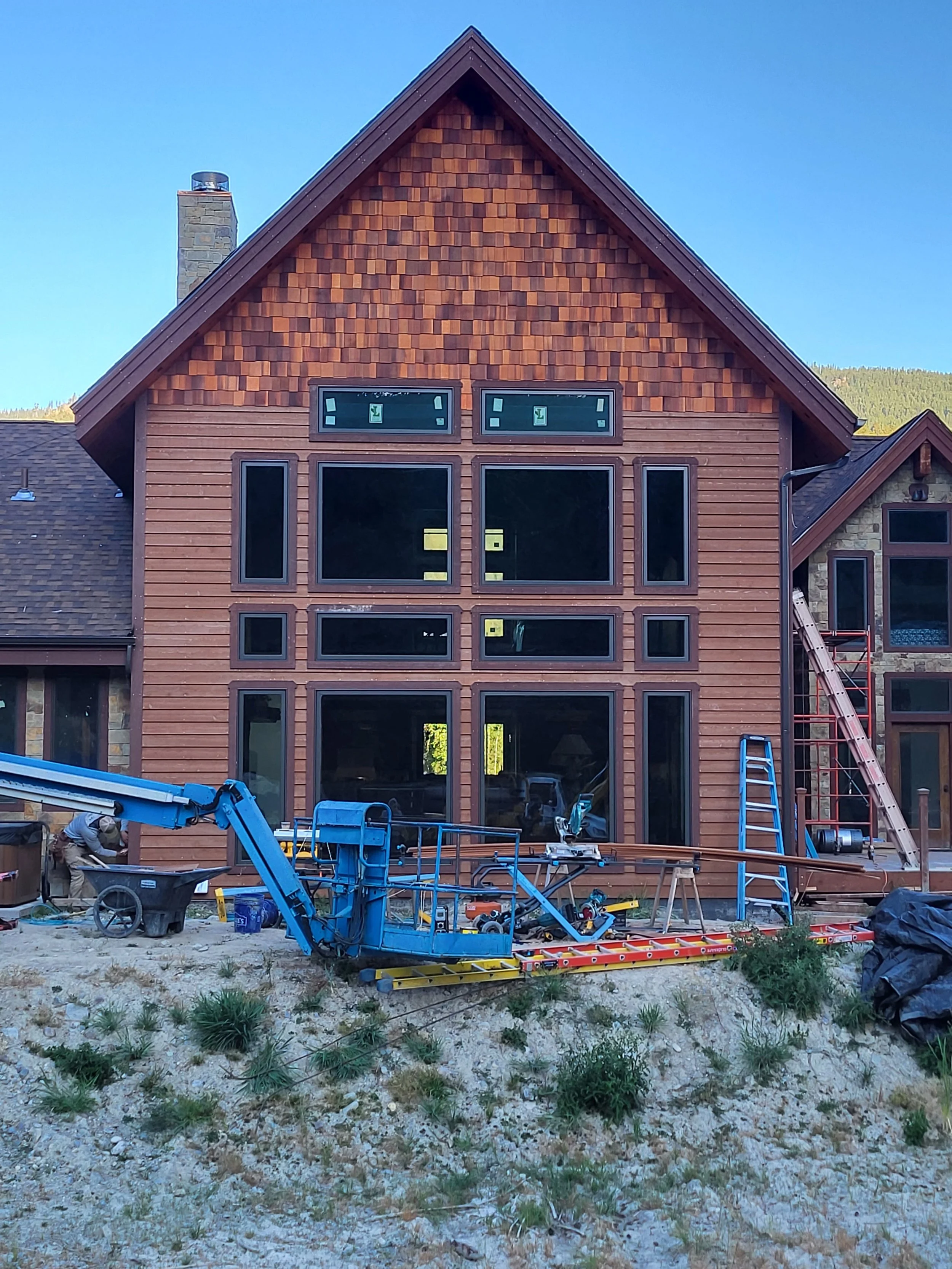 Construction of a large, multi-story house with wooden and stone exterior, several windows, and construction equipment and tools in the front yard.