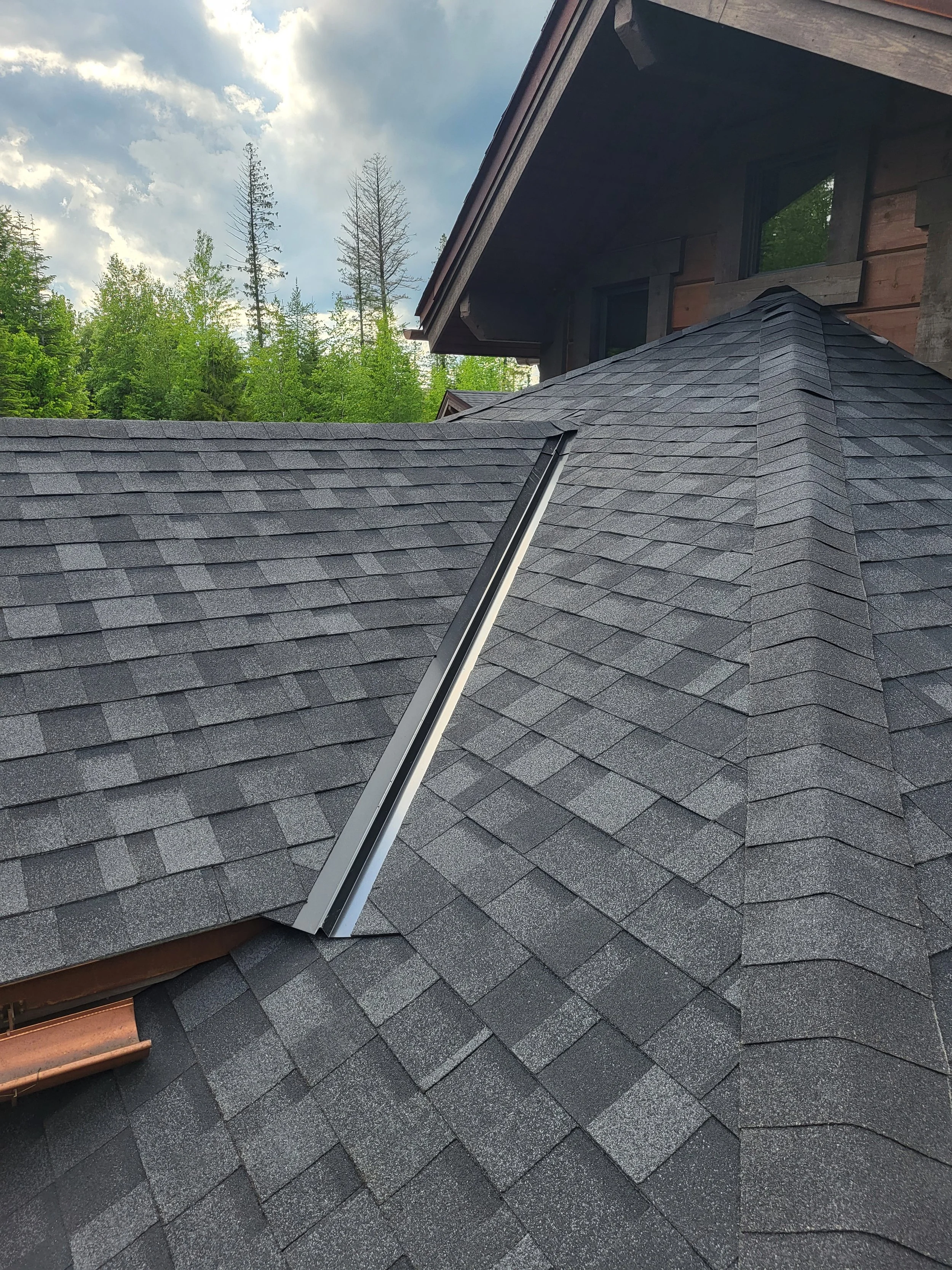 View of a house roof with asphalt shingles, a metal ridge vent, and trees in the background under a partly cloudy sky.