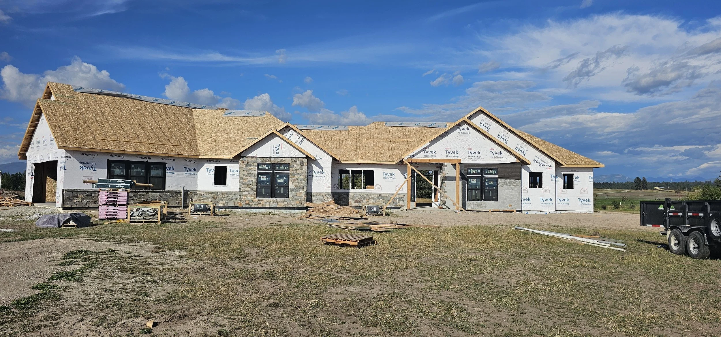 Under construction house with partially finished roof, stone exterior, black window frames, construction materials scattered around, on a dirt lot under a blue sky with clouds.