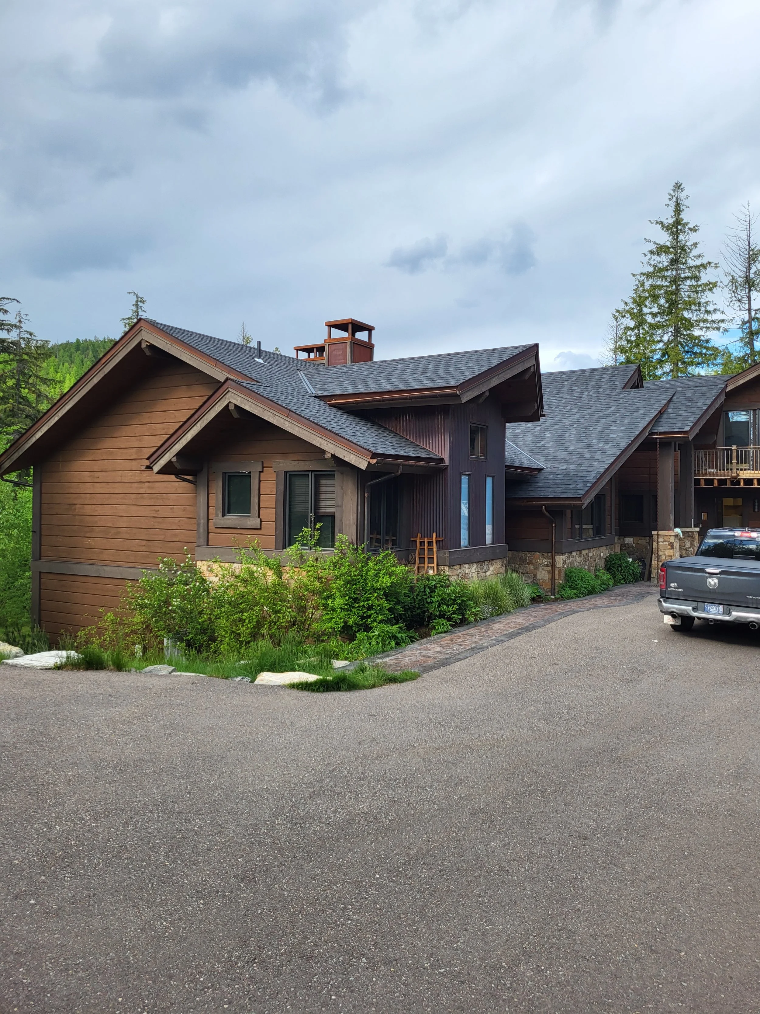 A large, modern house with brown wooden siding, a dark gray shingled roof, and multiple gables, located in a green, wooded area, with a paved driveway and a gray pickup truck parked outside.