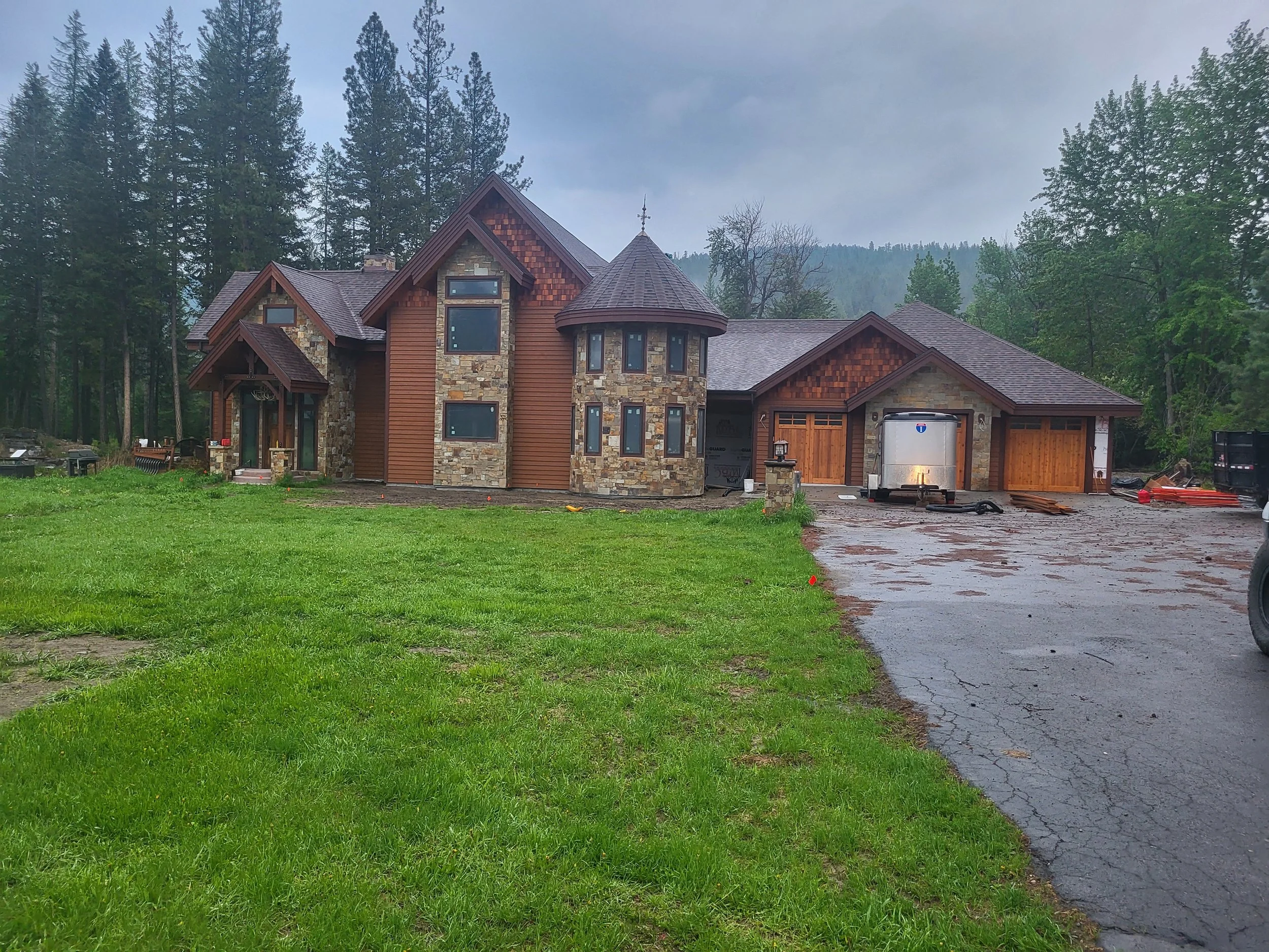 Large house under construction with stone and wood exterior, surrounded by greenery and tall trees, with a wet driveway and cloudy sky.