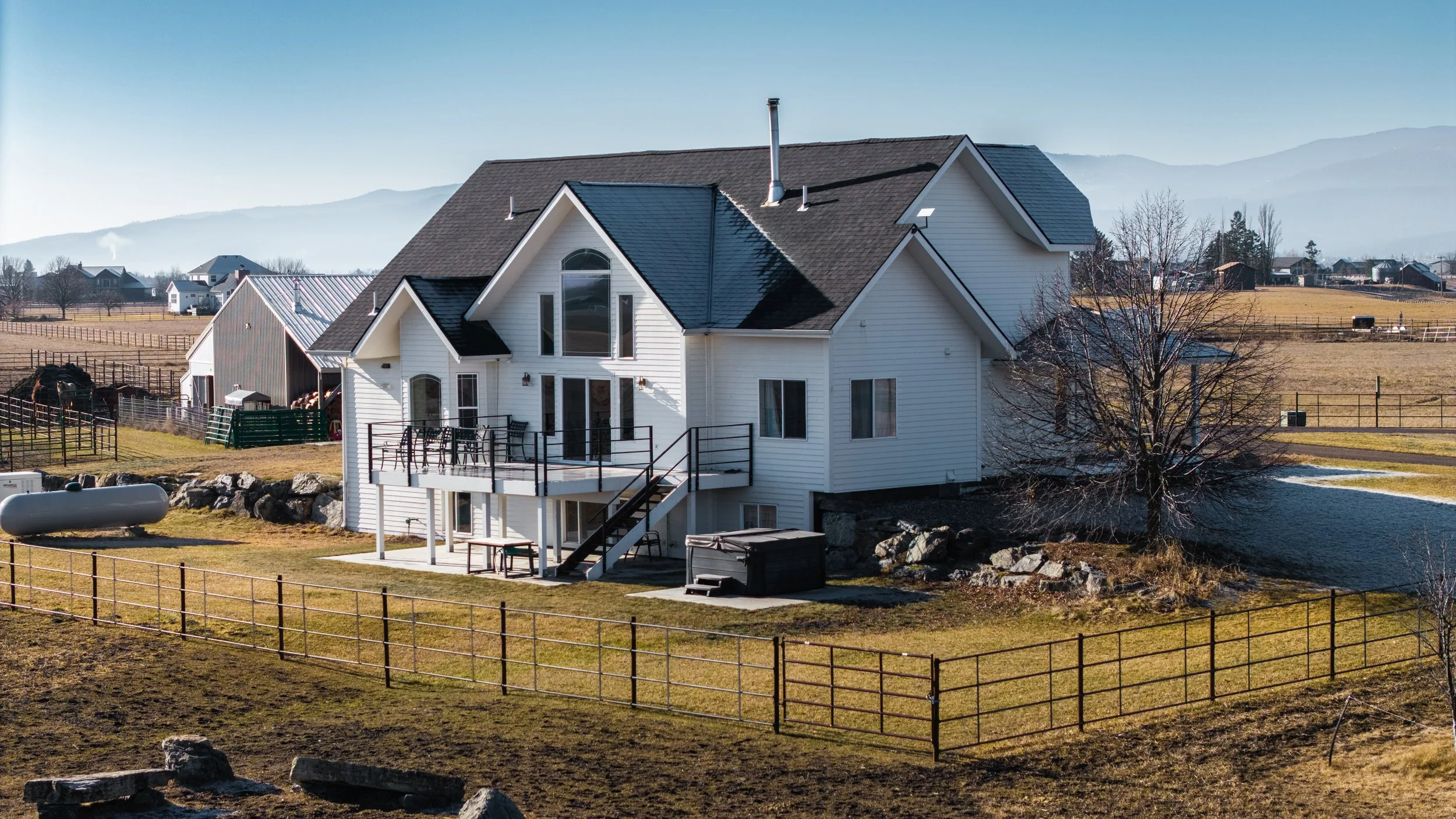 A large white house with multiple gabled roofs and a deck overlooking a fenced yard, with a pond and mountains in the background.