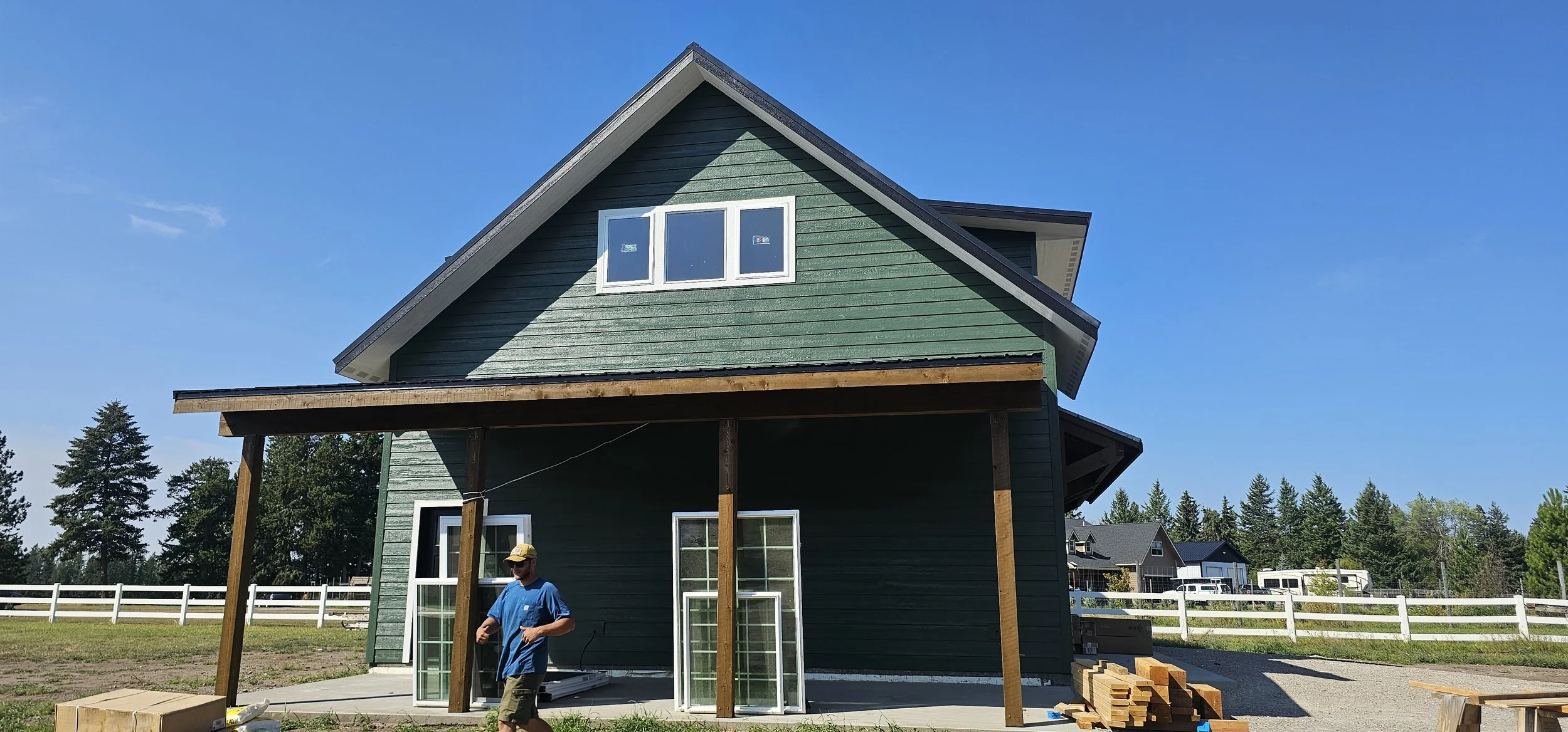 A two-story green house under construction with a man walking nearby, windows leaning against the house, and wood planks stacked on the ground, in a sunny yard with a white fence and other houses in the background.