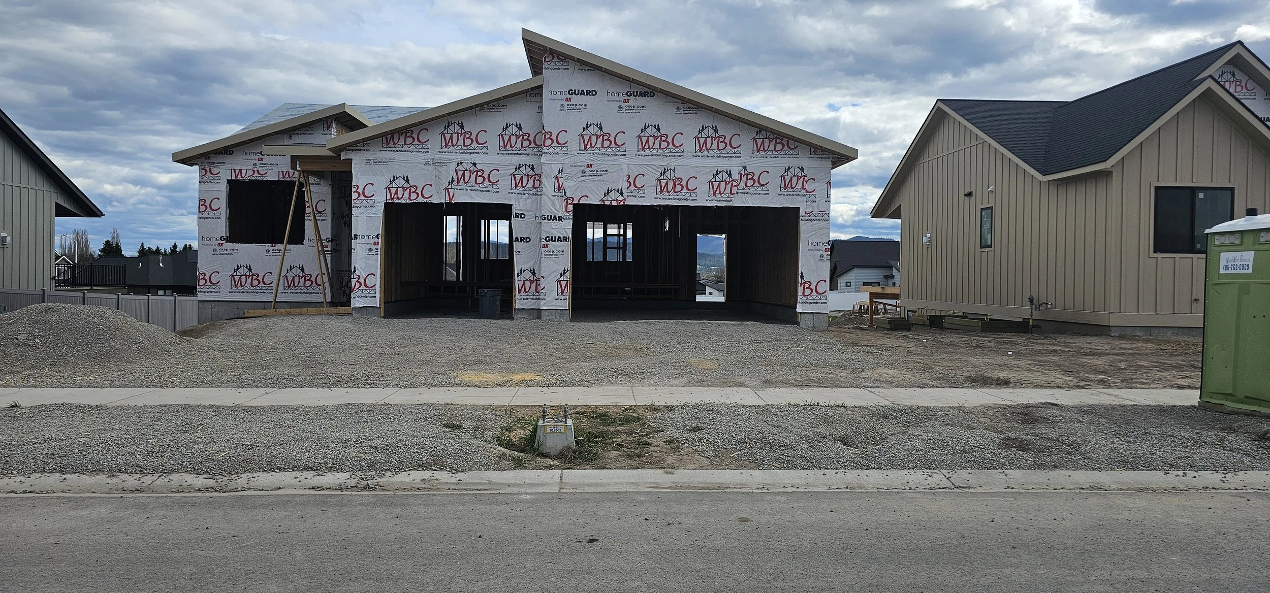 Front view of a house under construction with its frame exposed, wrapped in building wrap that has logos, surrounded by neighboring completed houses, with a cloudy sky overhead.