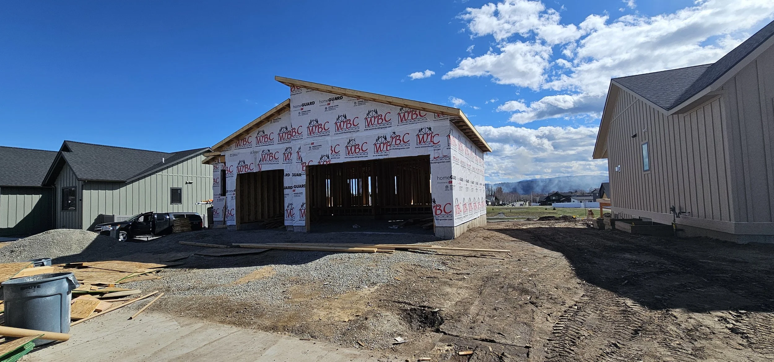 Under-construction house with framing and weather barrier, neighboring completed houses, gravel driveway, and construction materials on site under a blue sky with scattered clouds.