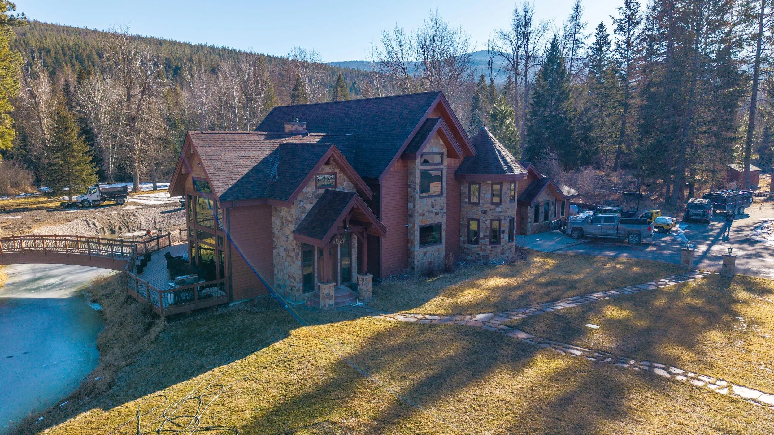 Large house in a forested area with a pond and a stone pathway leading to the front door. The house has a mix of stone and wood exterior, with multiple gables and windows, and surrounded by trees and parked trucks.