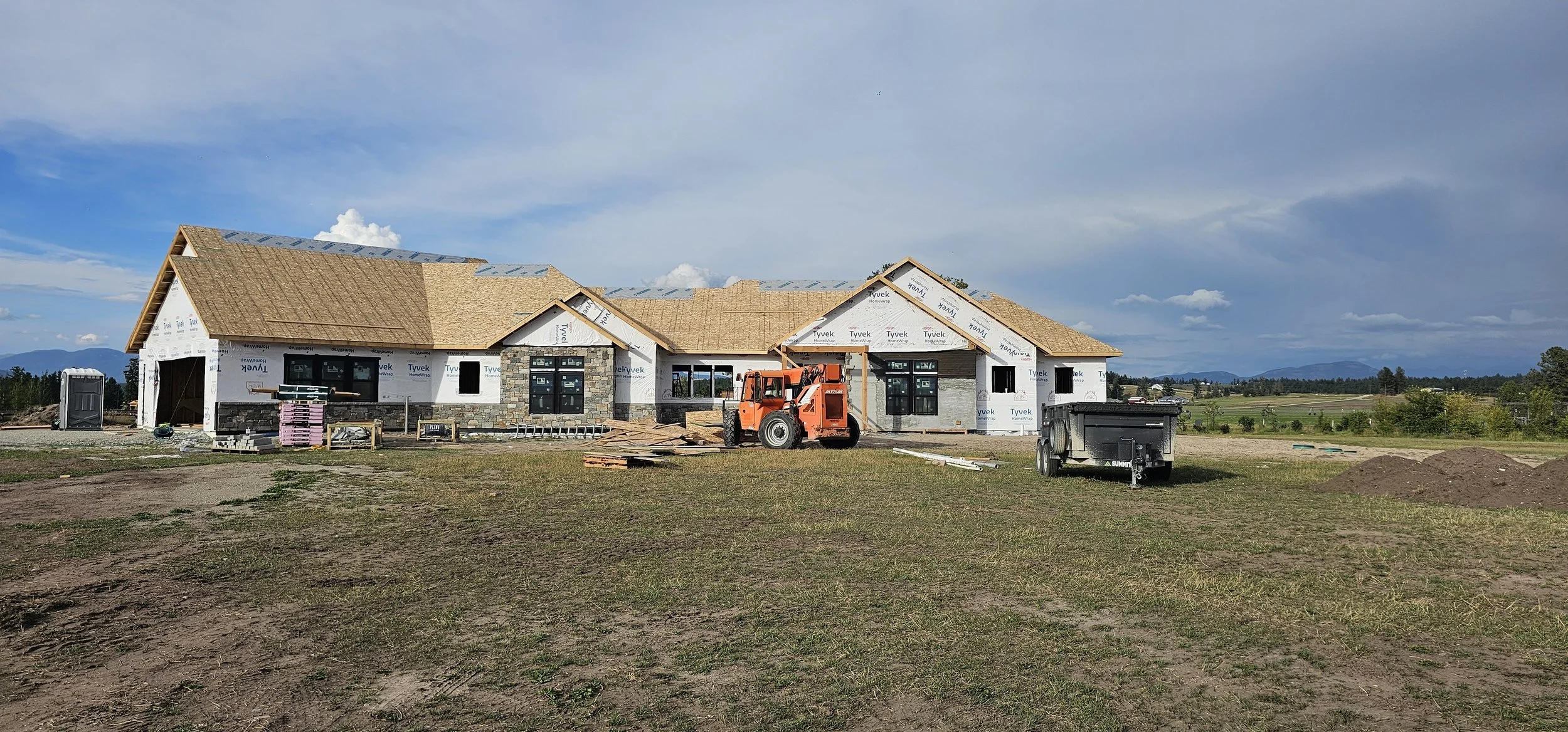 A house under construction on a large open lot with construction equipment, including a skid-steer loader and a trailer. The house has a partially finished roof and window installations.
