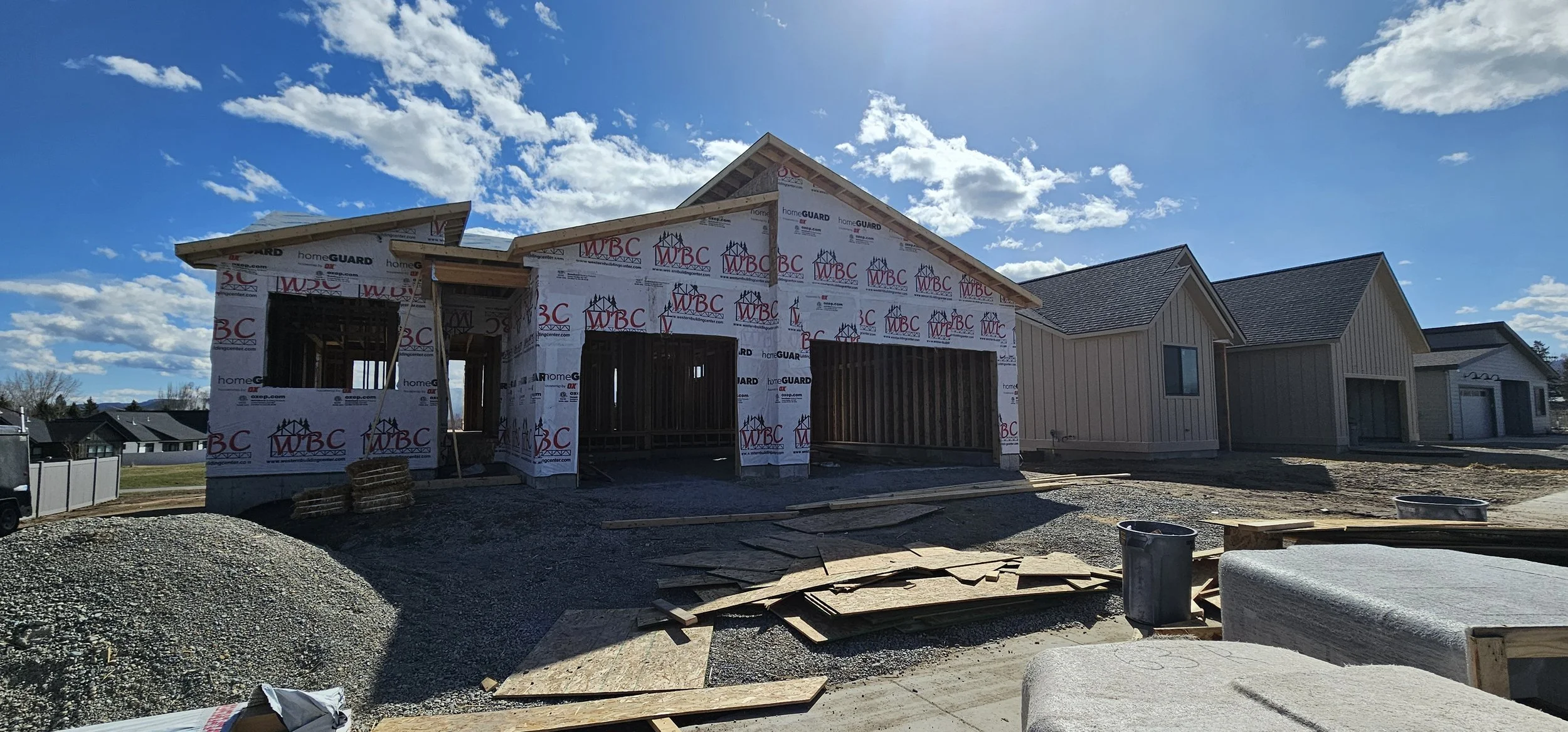 House under construction with framing and exterior sheathing, surrounded by construction materials and equipment, under a partly cloudy sky.