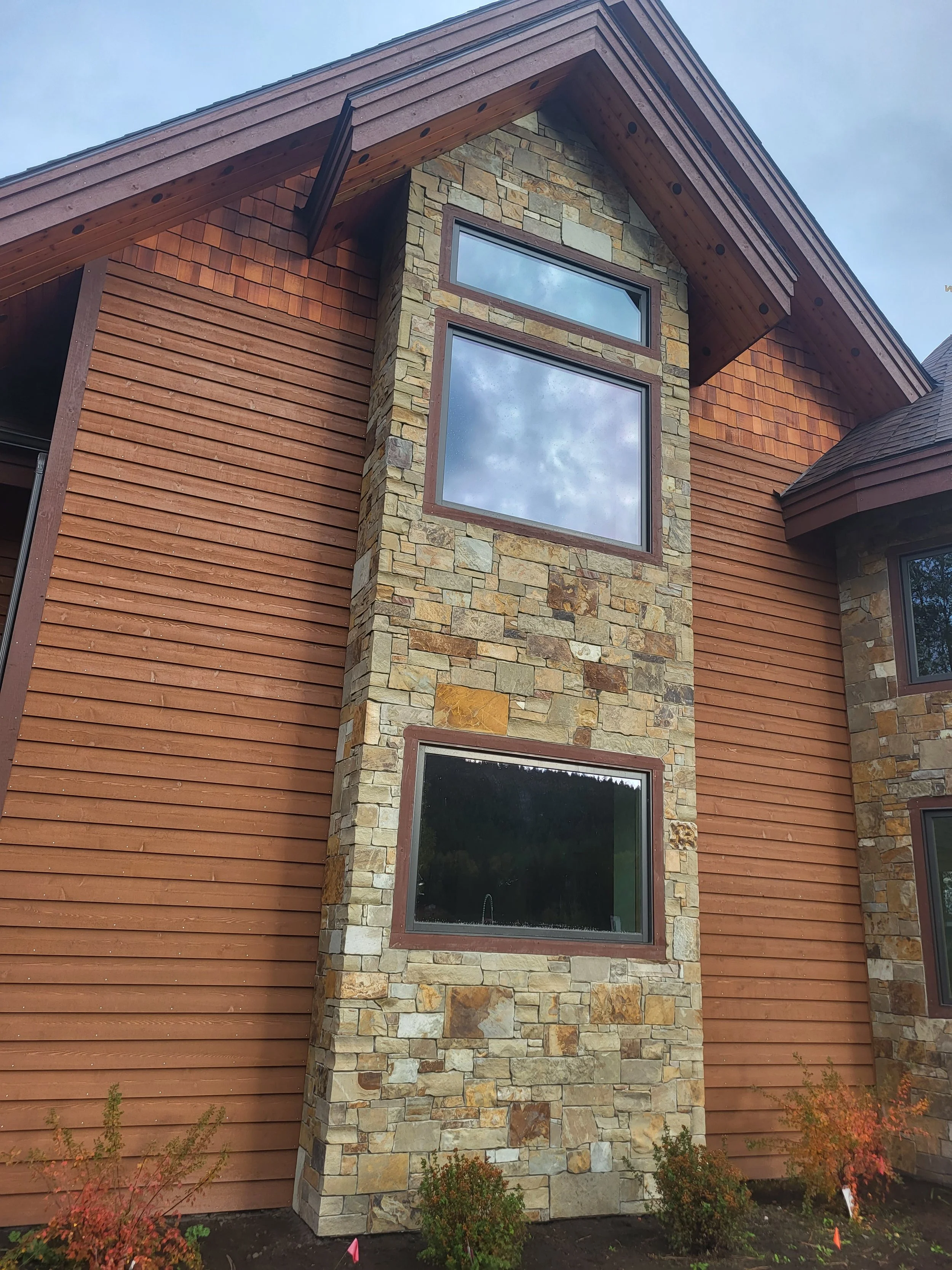 Close-up view of a house exterior featuring a stone chimney with three rectangular windows, brown wooden siding, and a brown roof with overhangs; small shrubs with some red leaves are planted at the base.