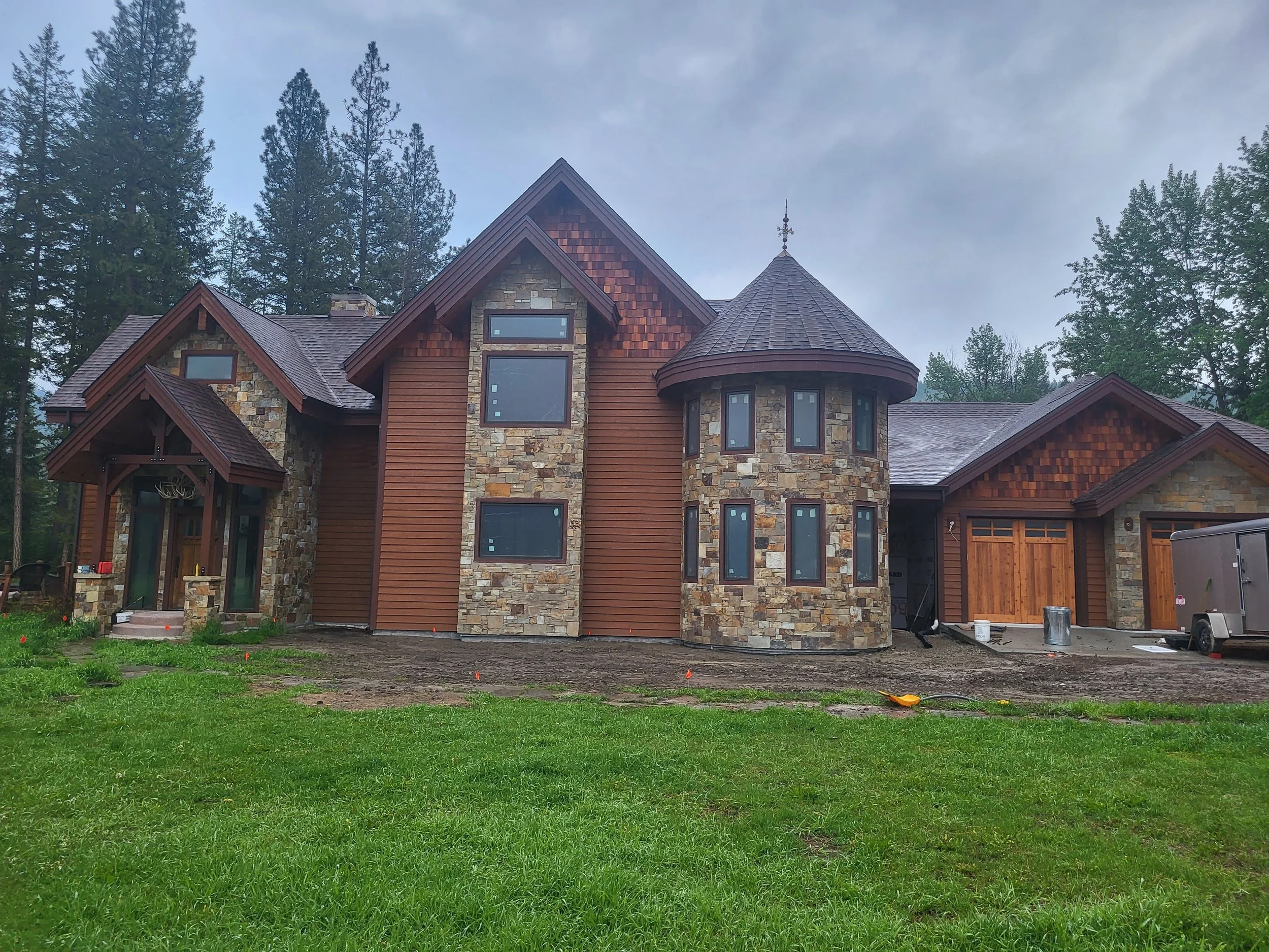 Front view of a large house under construction with a mix of stone and wood exterior, surrounded by trees with a grassy lawn in the foreground.