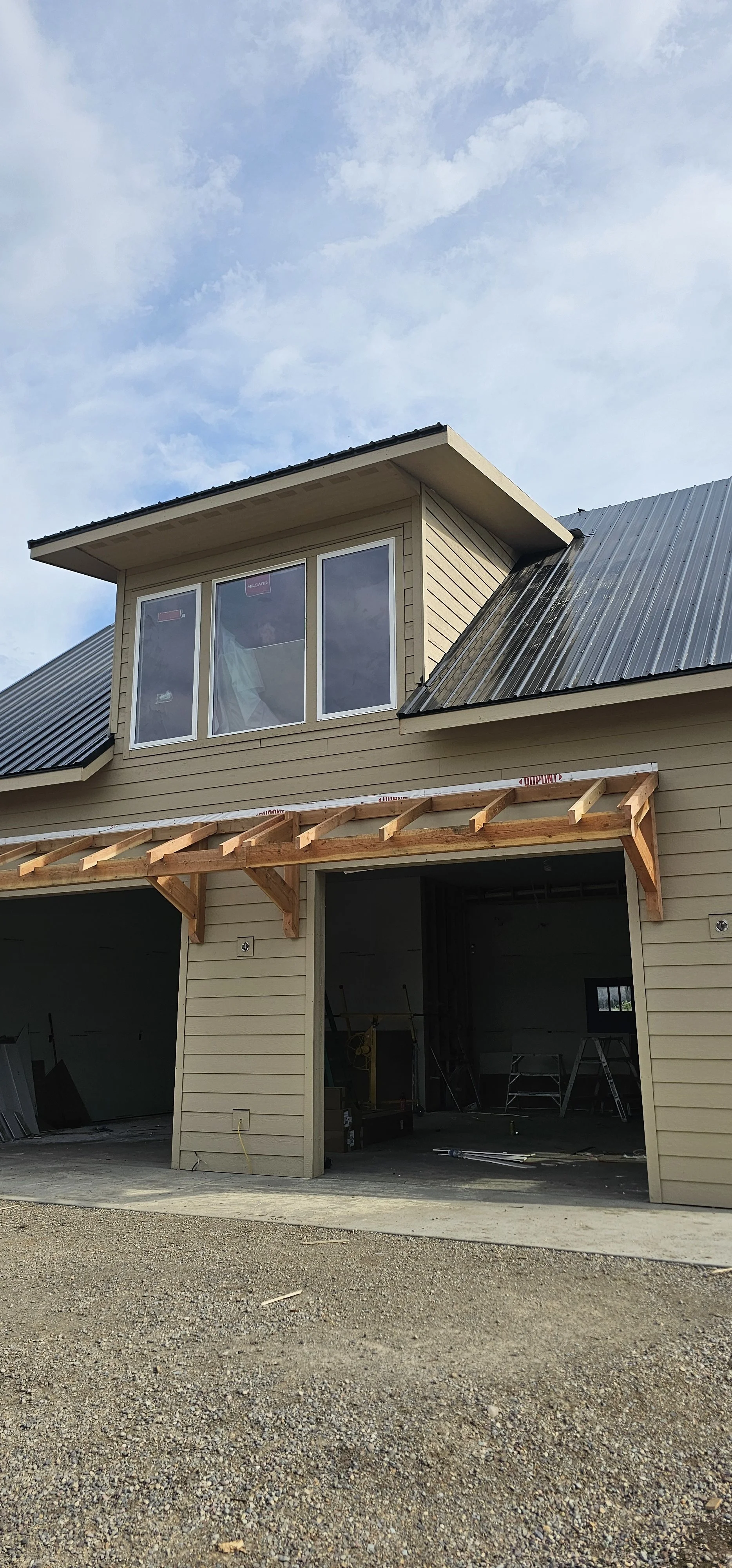 A house under construction with a two-story garage and an upper windowed room. The garage is open, revealing tools and ladders inside. The house has beige siding and a metal roof, with a wooden frame extending from the second floor indicating ongoing